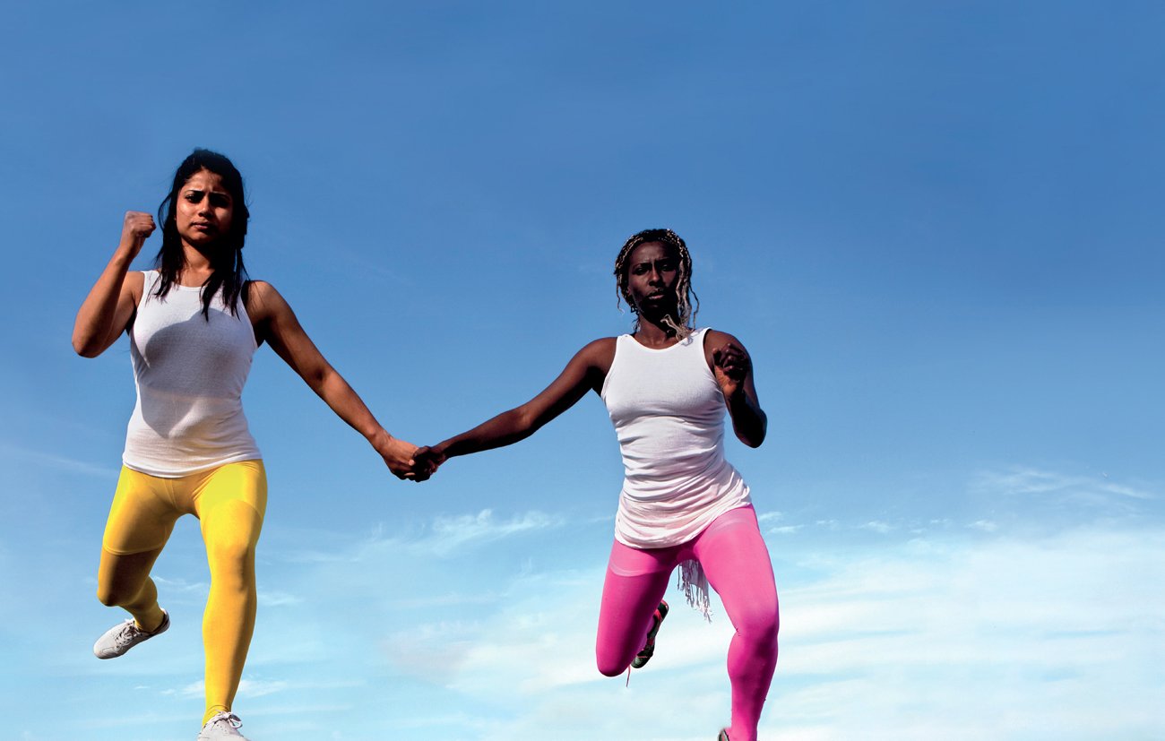 L'image montre deux femmes qui semblent flotter dans les airs, se tenant par la main. Elles portent des vêtements de sport, l'une en leggings jaunes et l'autre en leggings roses, avec des t-shirts blancs. En arrière-plan, on peut voir une ville urbaine s'étendre sous elles, avec un ciel bleu clair. Cette composition crée une sensation de liberté et de mouvement dynamique.