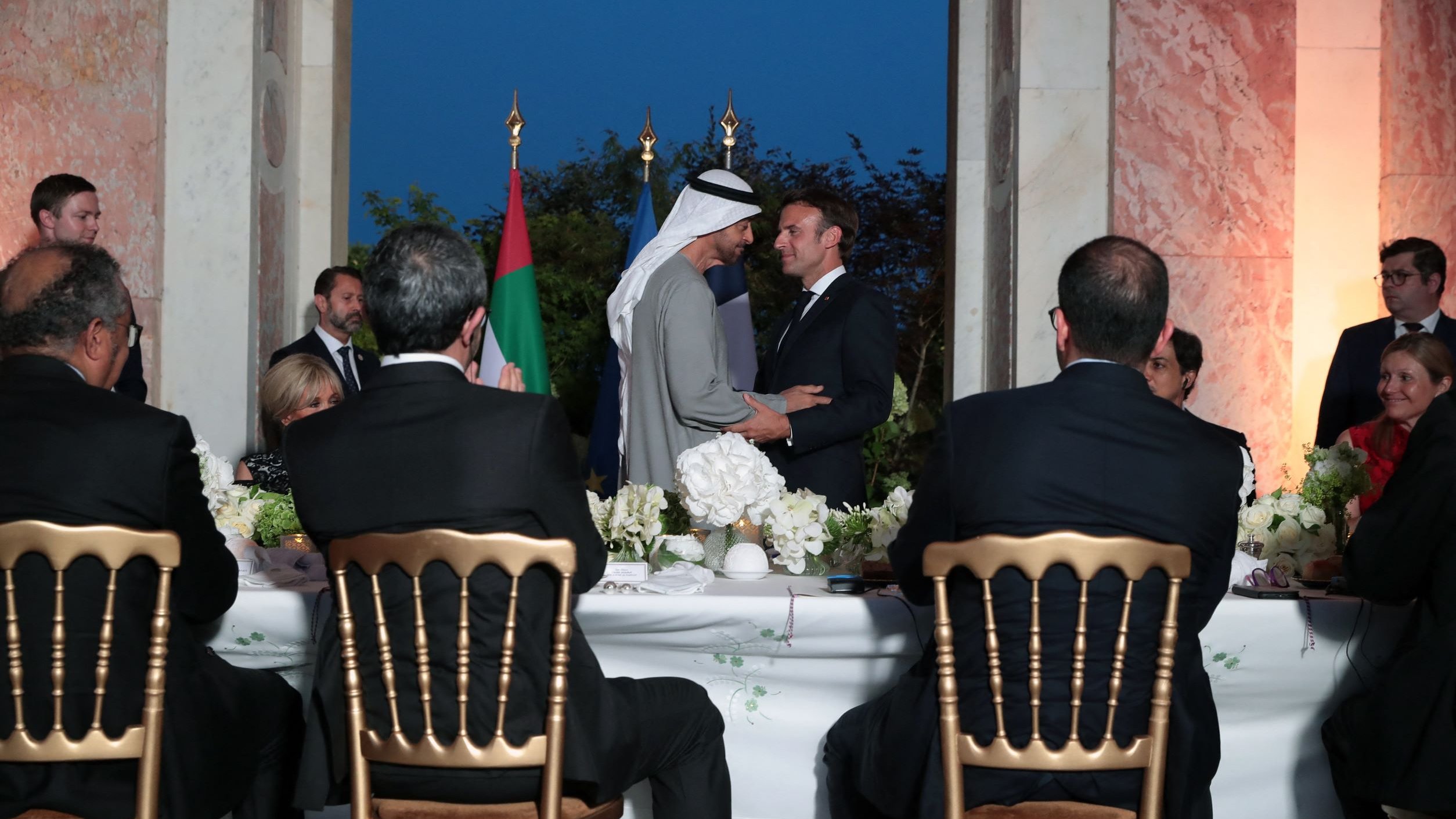 L'image montre une scène formelle dans un cadre élégant, probablement lors d'un dîner ou d'une réception. Au centre, deux hommes se serrent la main. On peut voir des drapeaux en arrière-plan, suggérant un contexte diplomatique. À la table, plusieurs personnes assistent à l'événement, avec une attention particulière sur les deux protagonistes. L'ambiance est chaleureuse et conviviale, marquée par des éléments de décoration soignés, comme des fleurs.