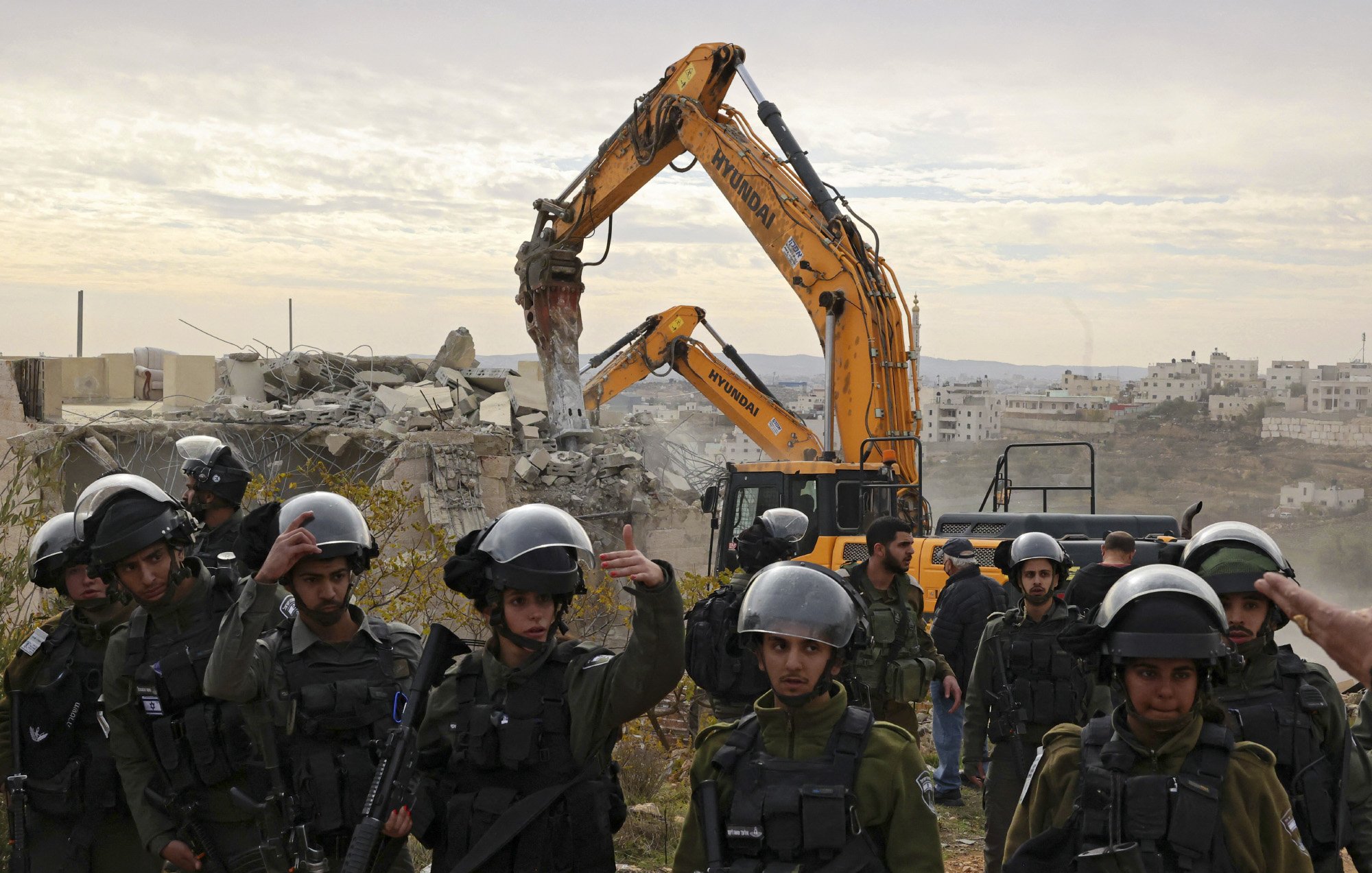 L'image montre une scène de destruction, où un bulldozer démolit un bâtiment en arrière-plan. Au premier plan, un groupe de soldats portant des casques et des équipements de protection surveille la situation. Ils semblent être en alerte, avec des armes en main. L'environnement est marqué par des décombres et un paysage urbain, suggérant un contexte de conflit. Le ciel est nuageux, ajoutant une atmosphère somber à la scène.