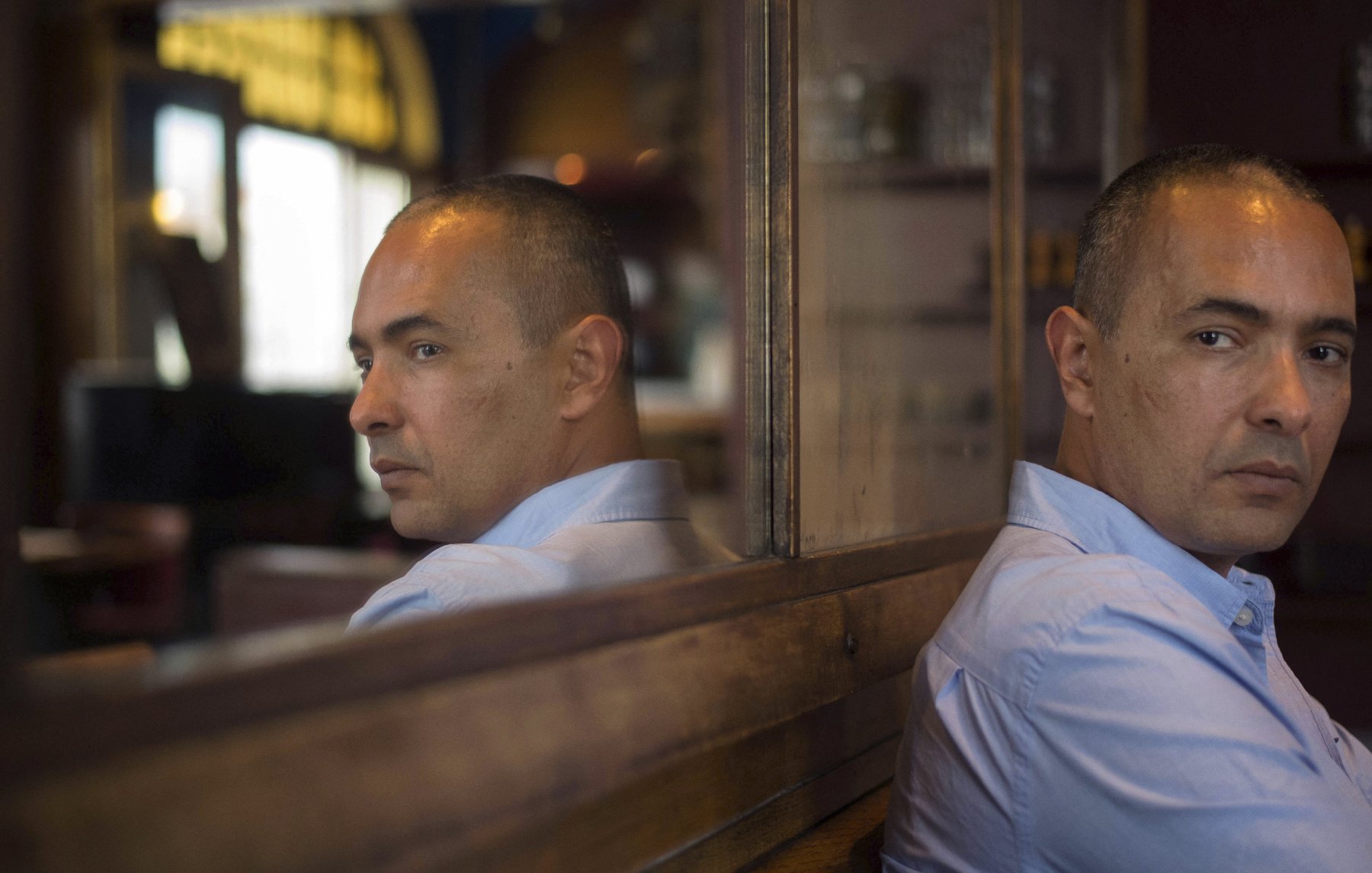 L'image montre un homme assis de dos, regardant vers un miroir qui reflète son visage. Il a une expression sérieuse, avec un léger froncement de sourcils. L'éclairage est doux et met en valeur les détails de son visage ainsi que l'ambiance de l'endroit où il se trouve, qui semble être un café ou un restaurant. Le décor en arrière-plan est flou, ce qui attire l'attention sur l'homme et son reflet.