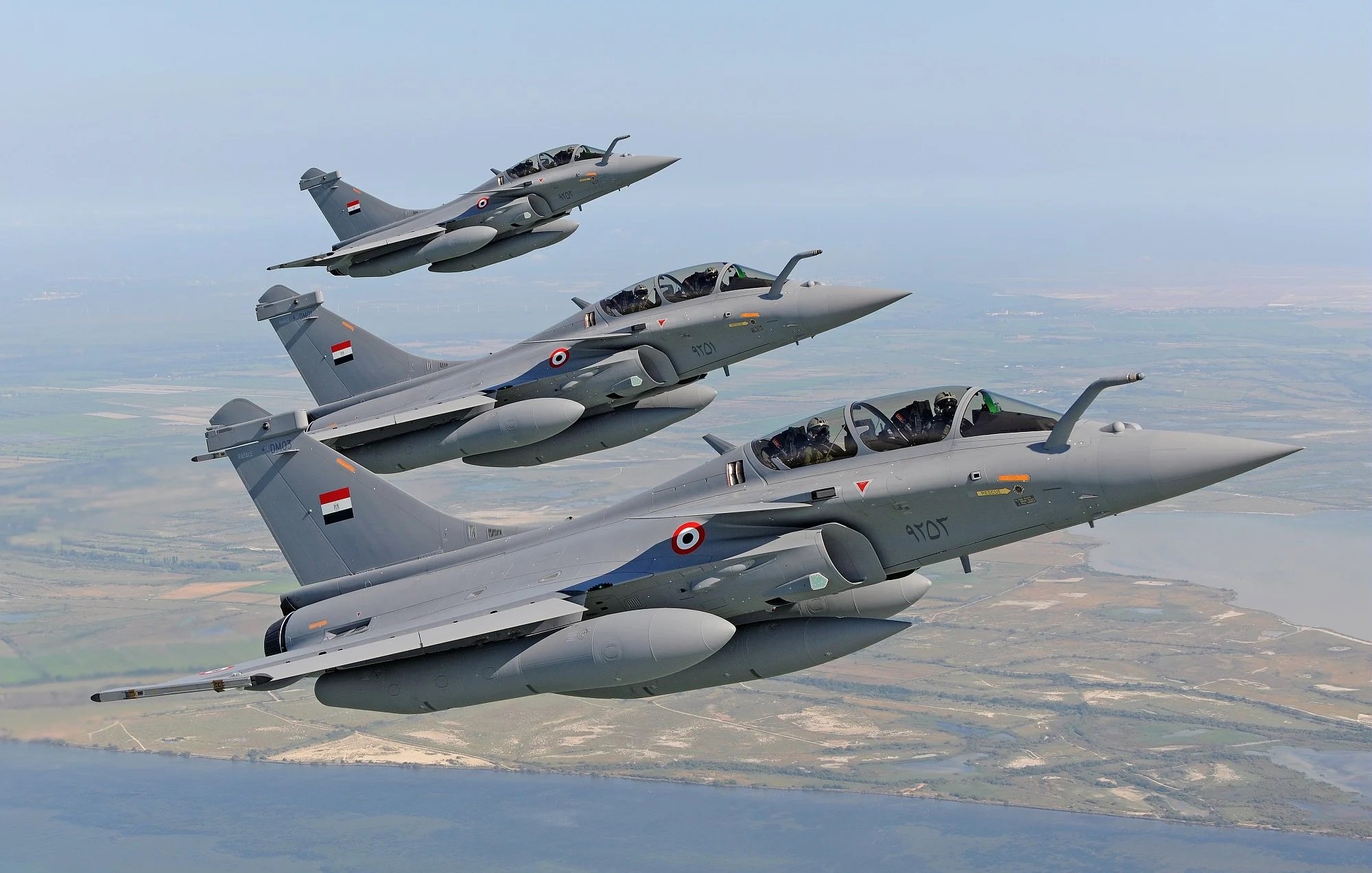 L'image montre un groupe de jets de chasse en plein vol. Trois avions de chasse, probablement des Rafales, sont alignés en formation, avec des couleurs principalement grises. Chaque avion porte des marqueurs et des emblèmes, semblant appartenir à une force aérienne. Le décor en arrière-plan indique un paysage vaste, avec des zones de terre et de l'eau visible, suggérant un vol à basse altitude. L'ensemble dégage une impression de puissance et de précision militaire.