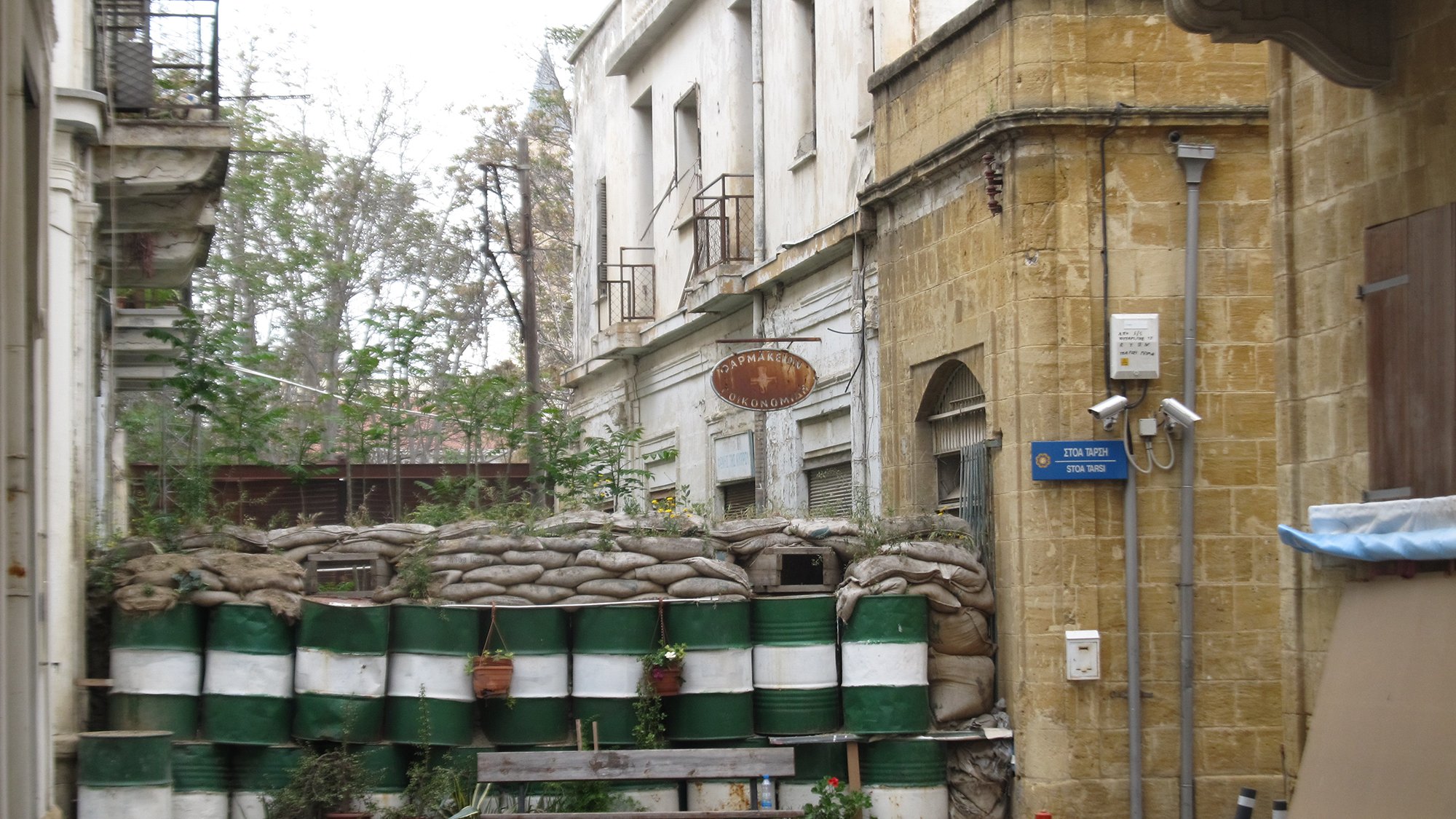L'image montre une rue étroite avec un mur constitué de sacs de sable et de fûts en plastique, probablement utilisés comme une barrière de protection. Au fond, il y a des bâtiments anciens avec des balcons, et des signes de végétation qui poussent sur le mur. L'atmosphère de la scène semble être à la fois urbaine et quelque peu abandonnée, avec un mélange de vie naturelle et d'éléments de construction.