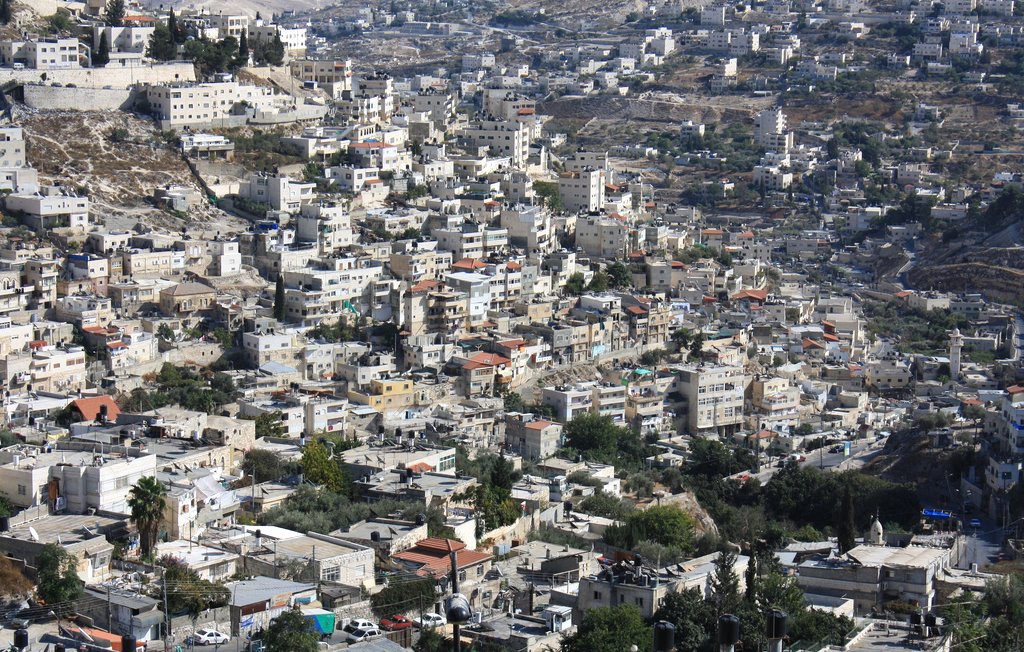 L'image montre un paysage urbain densément construit avec de nombreuses maisons et bâtiments. On peut observer une variété d'architecture, allant des structures modernes aux constructions plus anciennes. Les collines environnantes sont également visibles, avec des zones verdoyantes et des routes serpentant à travers le paysage. La ville semble être située dans une région montagneuse, avec un éclairage naturel qui met en valeur les détails des bâtiments.