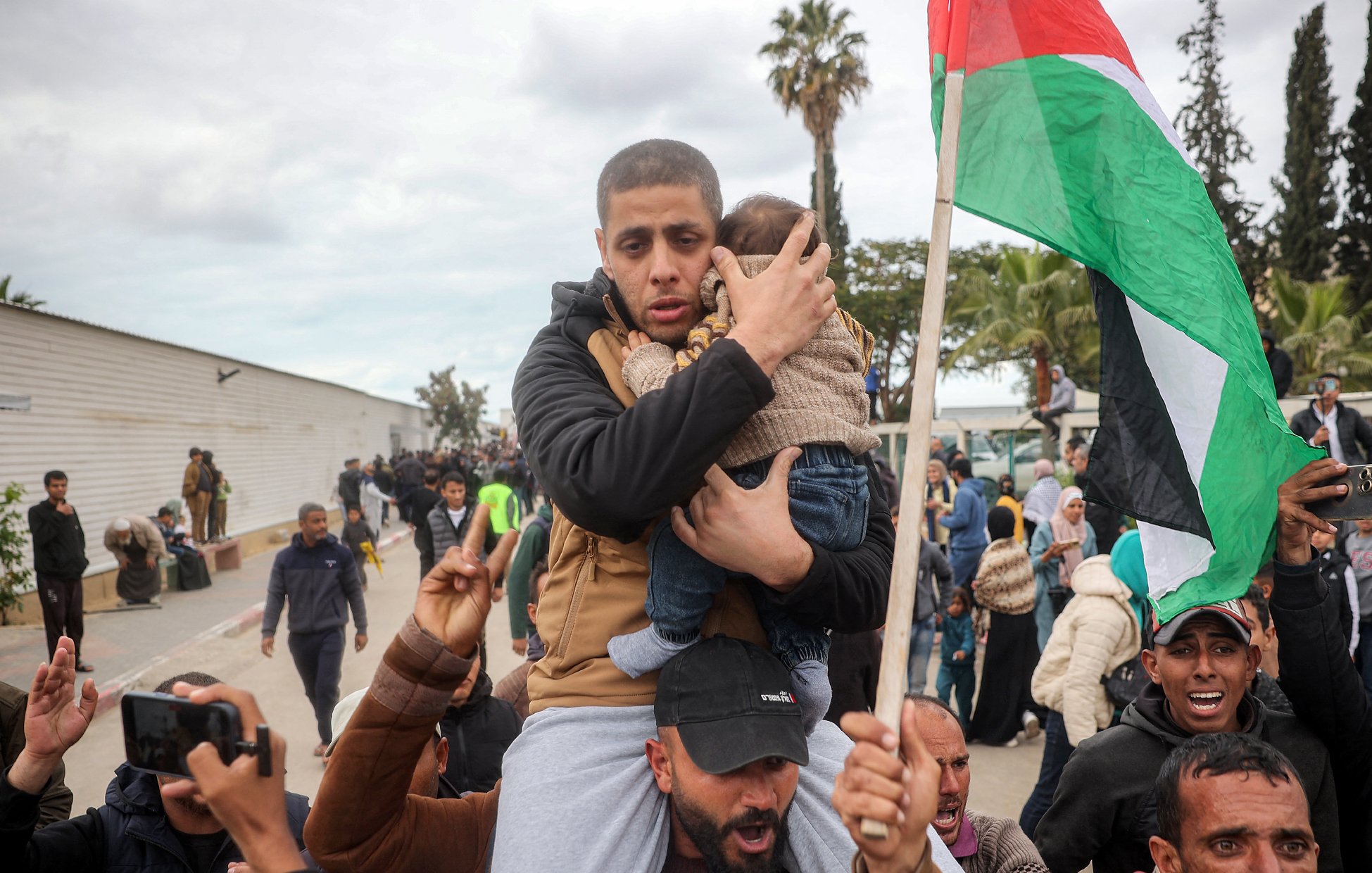 L'image montre une scène de rassemblement où un homme porte un enfant dans ses bras. Il semble vigilant et protecteur. En arrière-plan, il y a un groupe de personnes qui brandissent des drapeaux et expriment leur engagement ou leurs émotions. Des arbres et des structures sont visibles en arrière-plan, suggérant une ambiance de protestation ou de manifestation. L'atmosphère semble chargée en émotions.