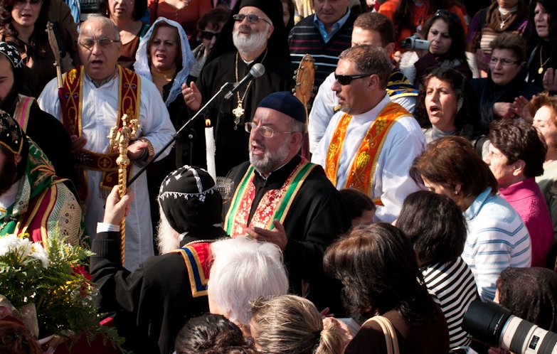 L'image représente un groupe de personnes rassemblées lors d'une cérémonie religieuse. Au centre, des figures cléricales en vêtements liturgiques colorés chantent et prient, entourées de fidèles. On aperçoit des chants, des bougies et des fleurs décoratives, ce qui suggère un événement festif ou sacré. L'ambiance est chaleureuse et communautaire, avec une multitude de visages exprimant la dévotion.