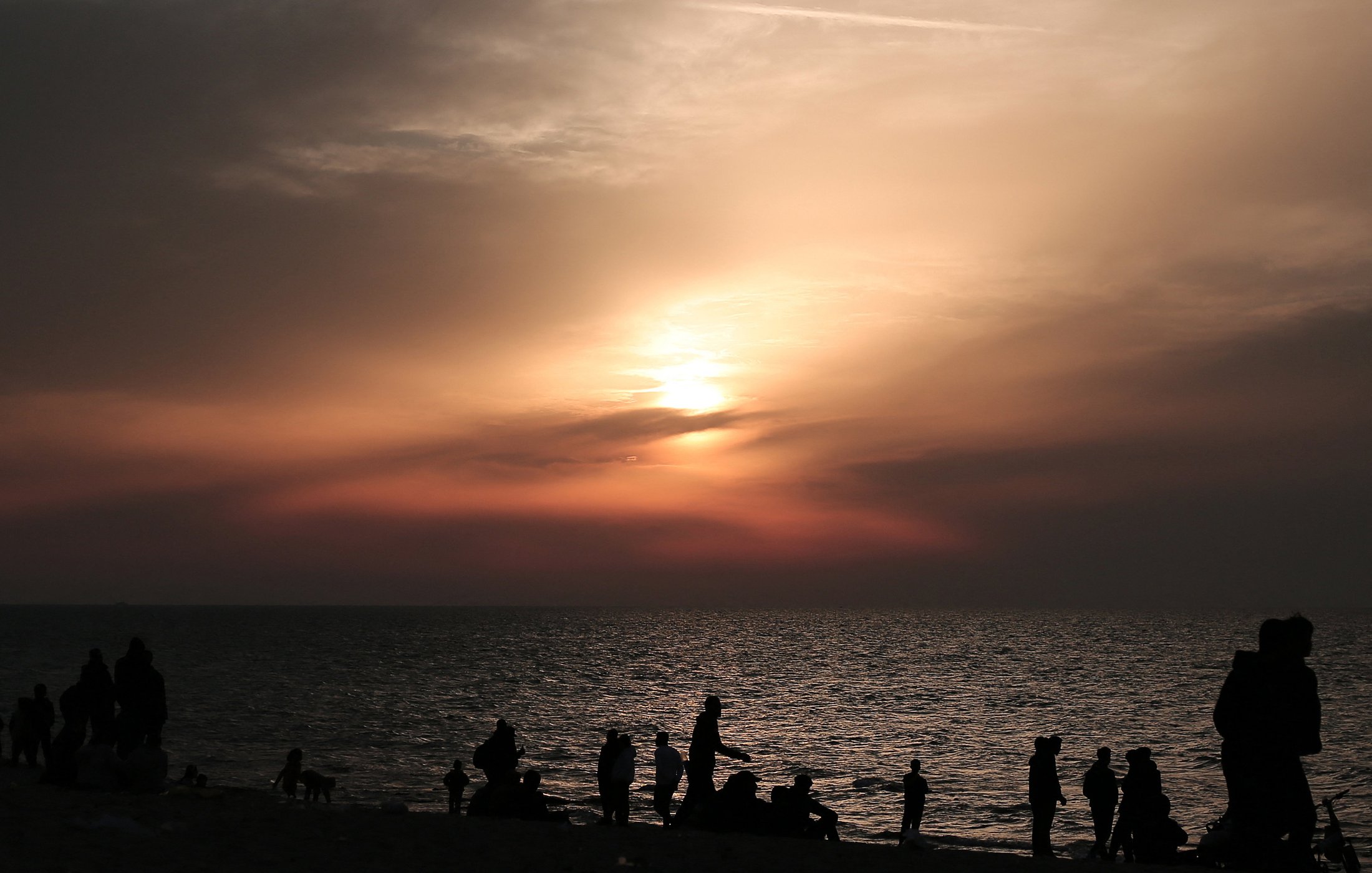 L'image montre une plage au crépuscule, avec des nuages qui tamisent la lumière du soleil couchant. De nombreuses silhouettes de personnes se déplacent le long du rivage, profitant de la scène. La mer reflète les couleurs douces du ciel, créant une atmosphère tranquille et paisible. Les silhouettes des individus ajoutent une dynamique à l'image, suggérant une ambiance conviviale et joyeuse.