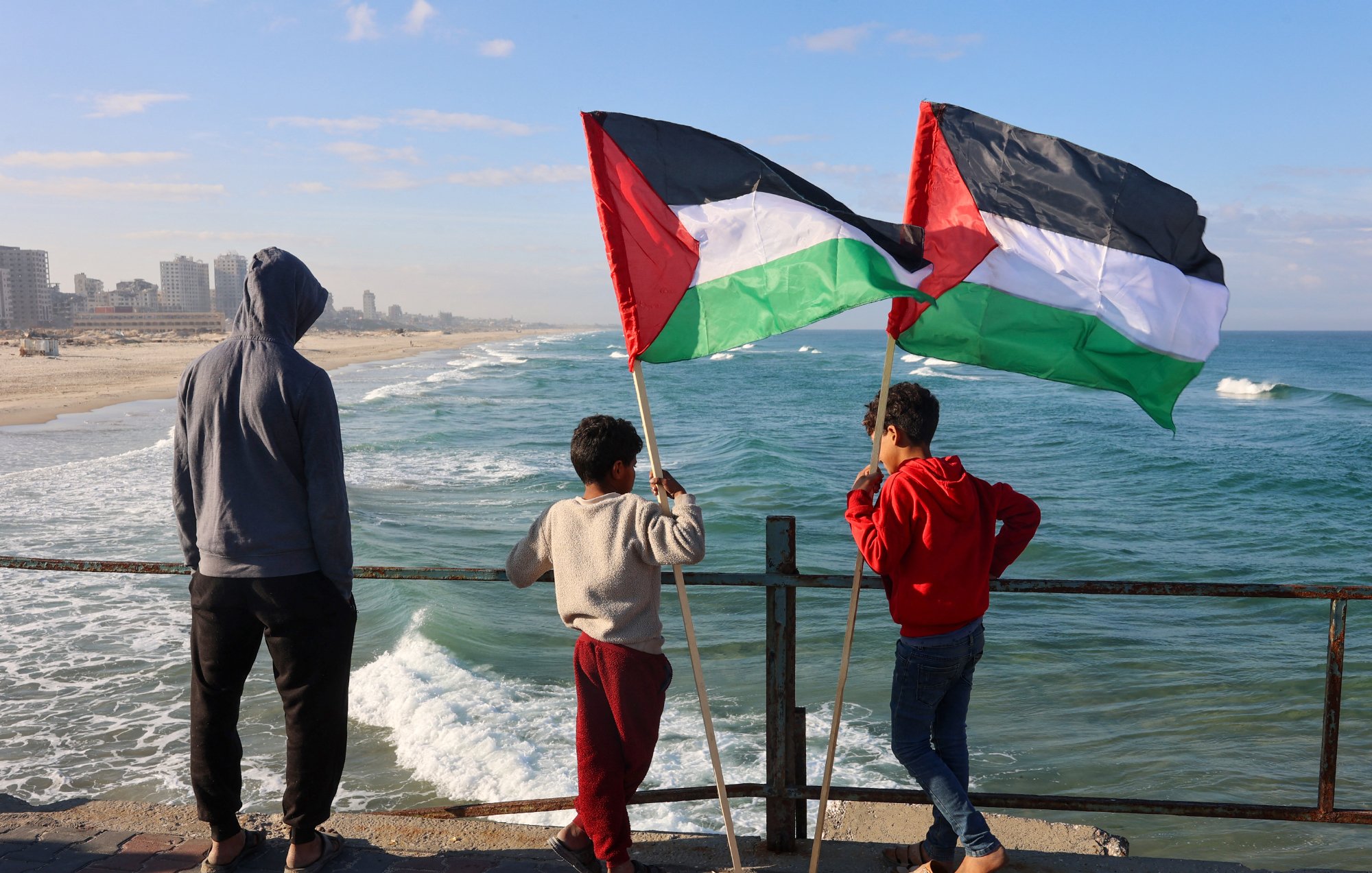 L'image montre trois enfants debout sur un bord de mer, regardant vers l'horizon. Deux des enfants tiennent des drapeaux avec les couleurs noir, rouge, vert et blanc, emblématiques de la Palestine. La mer est agitée, avec des vagues qui s'écrasent contre le rivage, et en arrière-plan, on peut apercevoir des bâtiments sur la plage. Les enfants semblent contemplatifs, profitant de l'instant. Le ciel est partiellement nuageux, créant une atmosphère tranquille.