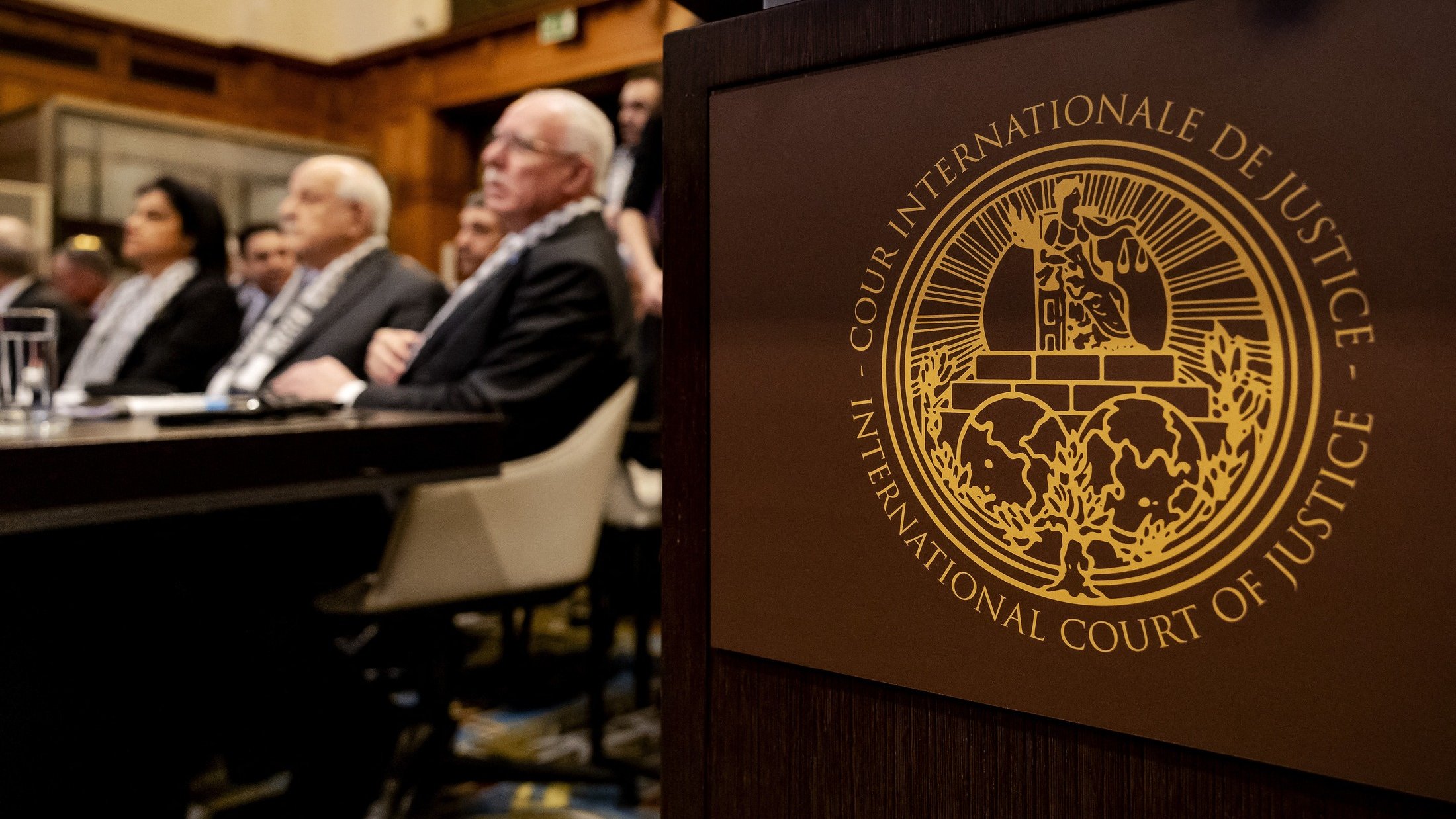 L'image montre une scène à la Cour internationale de justice. Au premier plan, on peut voir le symbole de la Cour, qui est un emblème distinctif. En arrière-plan, plusieurs personnes sont assises, probablement des membres du tribunal ou des délégués, attendant une session ou un discours. L'environnement semble formel, avec une décoration typique d'une institution judiciaire.