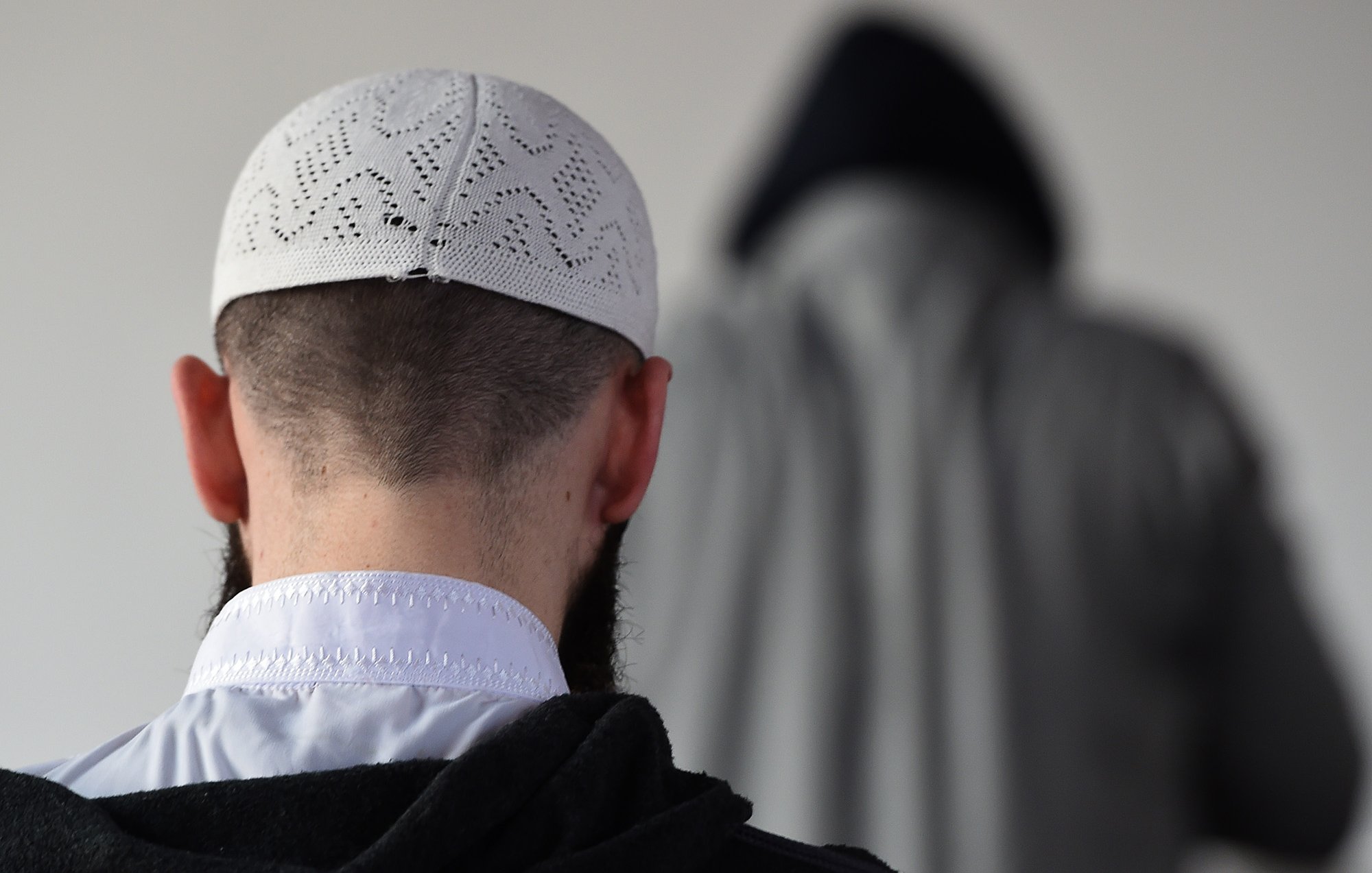 L'image montre un homme de dos, portant un taqiyah (un couvre-chef traditionnel), avec une barbe visible. En arrière-plan, on peut apercevoir une silhouette floue d'une autre personne, vêtue d'une cape ou d'un vêtement similaire. L'environnement semble simple et neutre, avec peu d'éléments distrayants. La scène évoque une ambiance calme et contemplative.