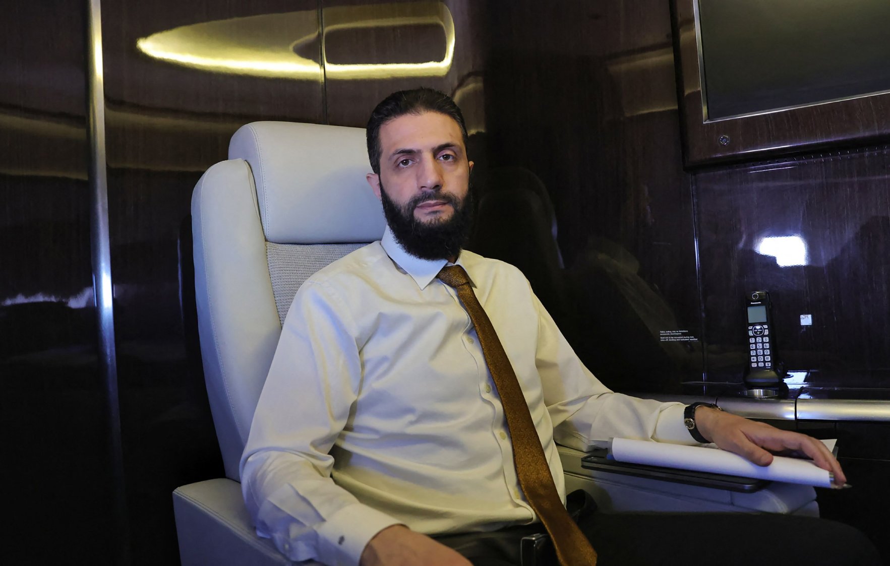 Un homme est assis dans un fauteuil confortable à l'intérieur d'un avion privé. Il porte une chemise claire et une cravate. L'environnement est élégant, avec des surfaces sombres et brillantes.