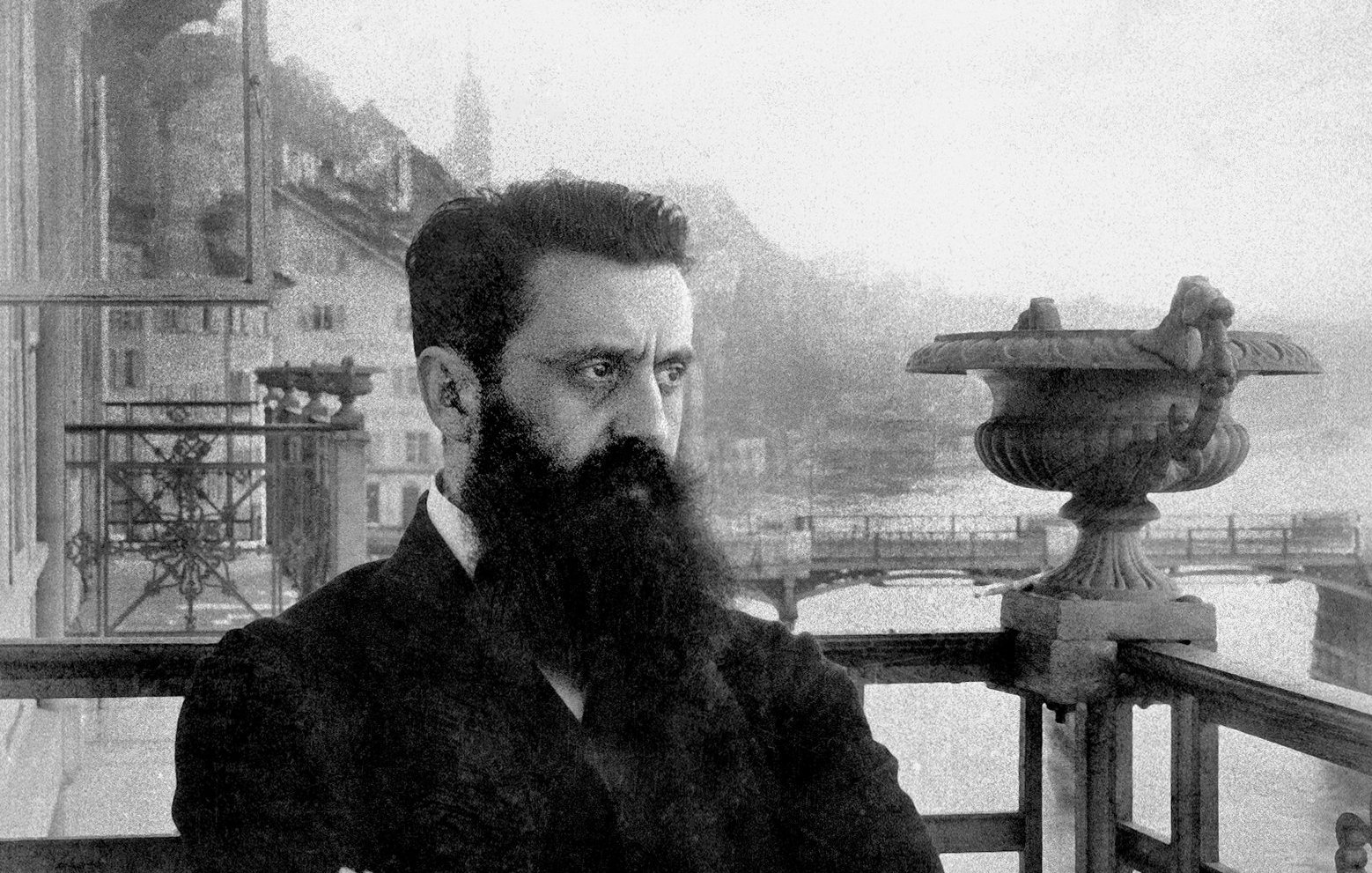 L'image montre un homme avec une barbe fournie, portant un costume sombre. Il est debout sur un balcon orné de balustrades métalliques, avec un regard pensif et sérieux. En arrière-plan, on aperçoit des montagnes et un plan d'eau, suggérant un paysage pittoresque. L'atmosphère est nostalgique, renforcée par le ton en noir et blanc de la photographie, ajoutant une dimension historique à la scène.