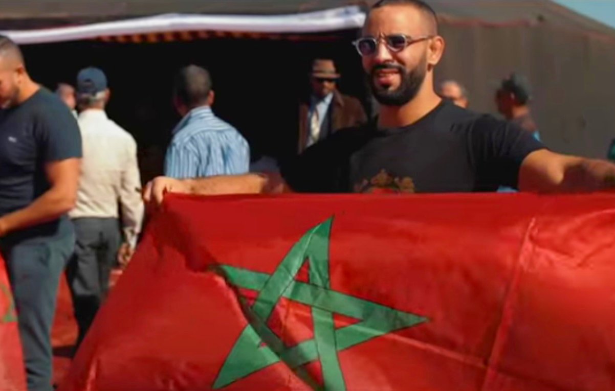 L'image montre un groupe de personnes, dont un homme tenant un drapeau marocain. Il sourit et affiche fièrement le drapeau rouge avec une étoile à cinq branches verte. En arrière-plan, on aperçoit d'autres personnes et des tentes, ce qui laisse supposer une rencontre ou un événement en plein air. L'atmosphère semble festive et conviviale, évoquant un rassemblement culturel ou patriotique.