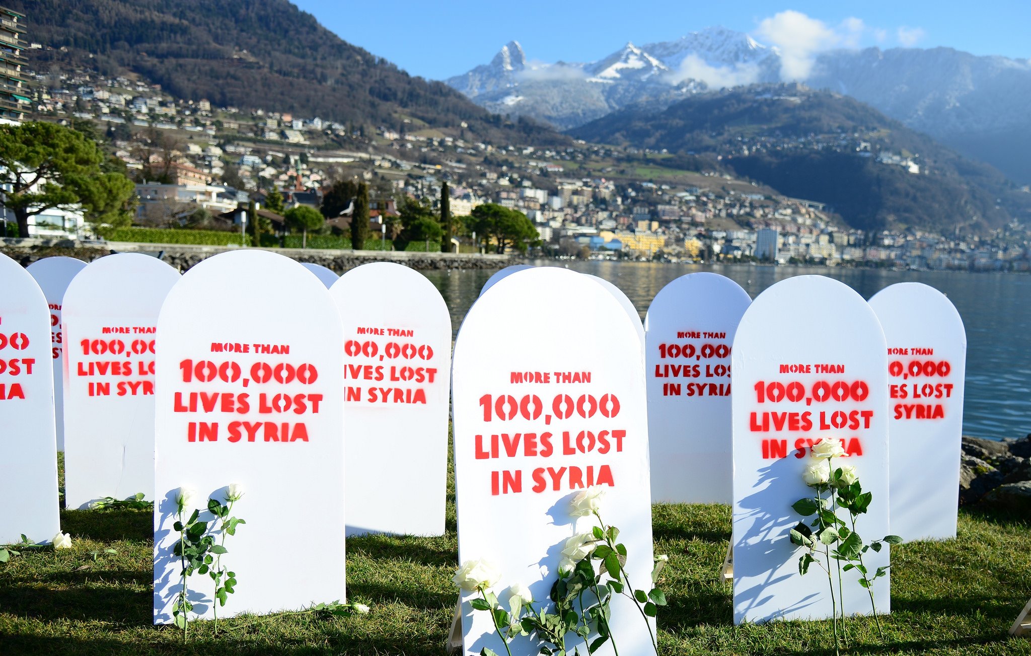L'image montre un rassemblement commémoratif avec plusieurs pierres tombales blanches disposées sur un terrain vert. Chaque pierre tombale est marquée de rouge avec des inscriptions telles que "More than 100,000 lives lost in Syria". À côté des pierres tombales, il y a des roses ou des plantes, symbolisant probablement la mémoire des vies perdues. En arrière-plan, on peut voir une vue magnifique de montagnes et d'une ville au bord de l'eau. L'ambiance générale semble être solennelle et réfléchissante, soulignant une tragédie humanitaire.