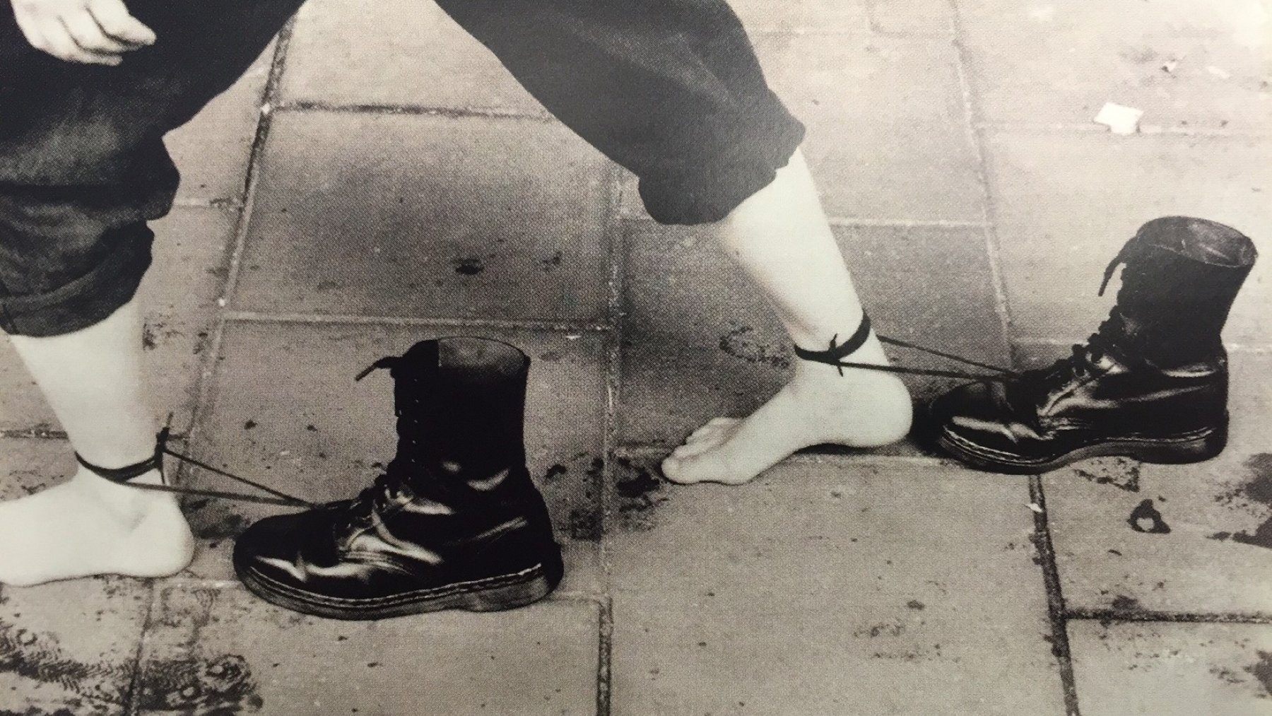 Des pieds nus liés à des bottes noires par des cordes. Image en noir et blanc.