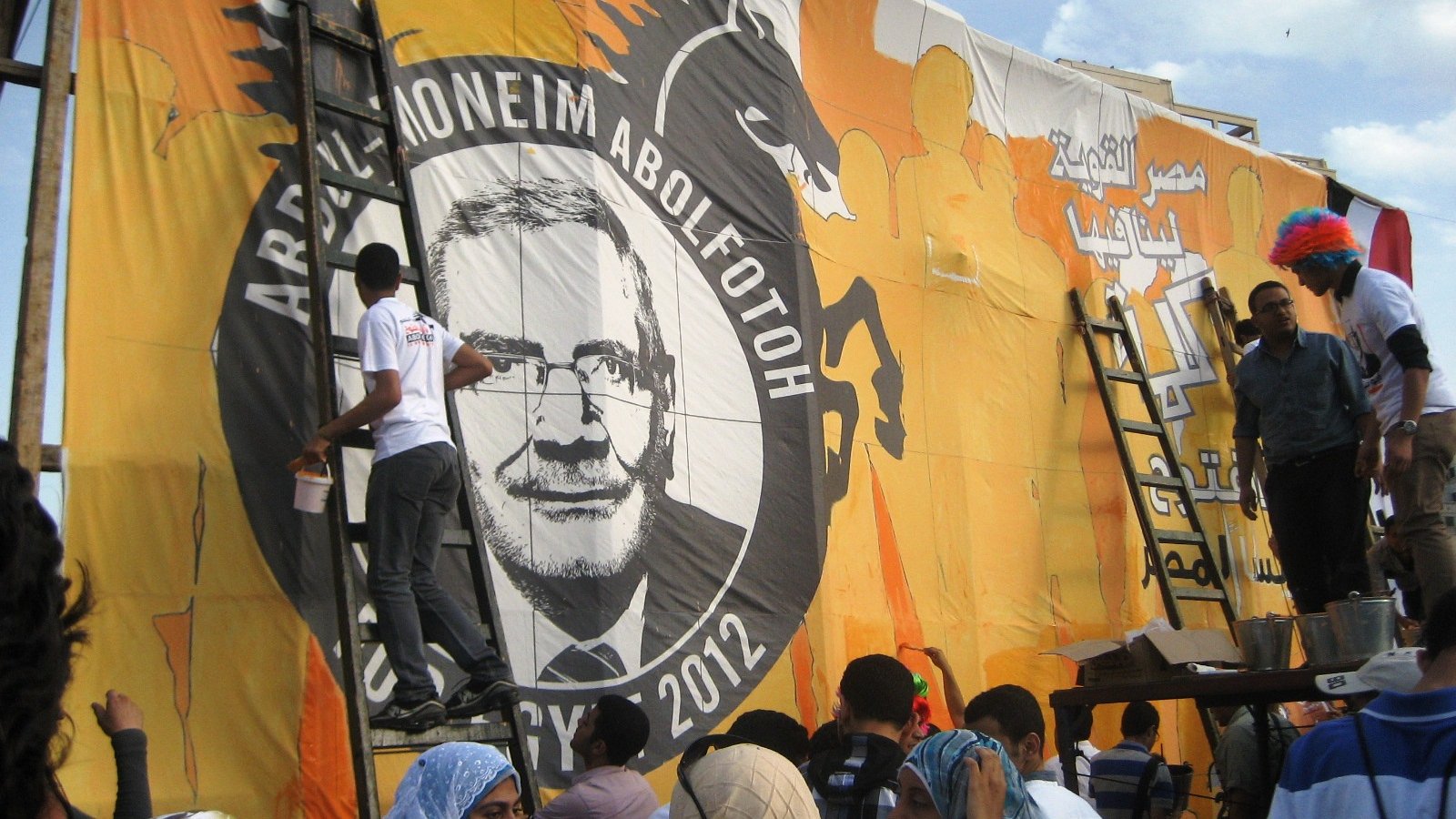 L'image montre un groupe de personnes en train de peindre une grande bannière ou une affiche. Ils sont sur des échelles et travaillent à mettre en place un portrait ou un dessin qui semble représenter une figure importante, peut-être liée à un événement politique. Le fond est coloré, principalement en orange, et il y a des inscriptions en arabe. Les femmes au premier plan portent des foulards, et l'atmosphère semble dynamique, avec des gens engagés dans le travail collectif.