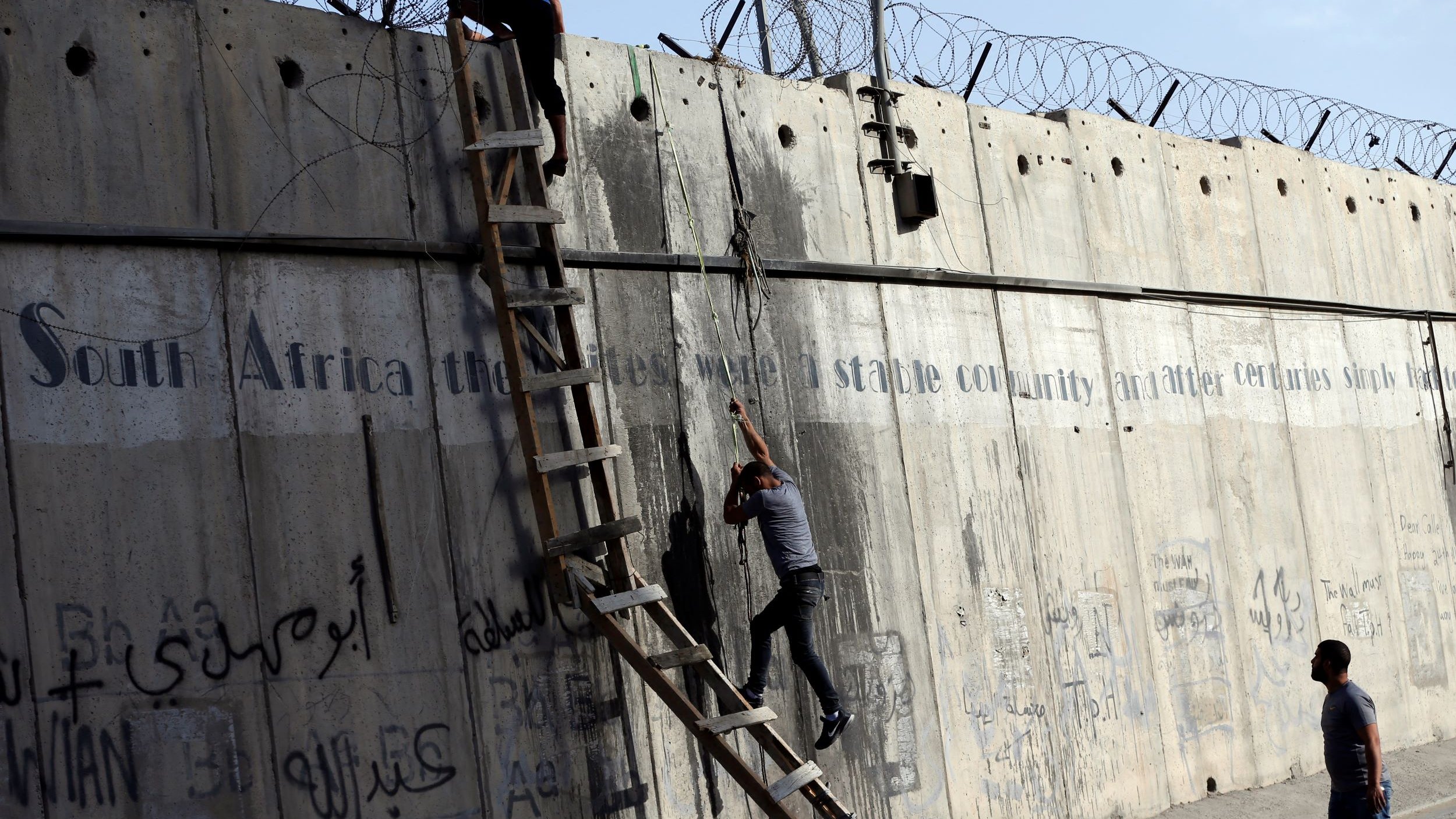 L'image montre une scène où deux personnes tentent de monter ou de descendre d'un mur de béton. Une échelle est appuyée contre le mur, et il y a des barbelés au sommet. Le mur présente également des graffitis ou des inscriptions, et l'environnement semble urbain. Les personnes semblent concentrées sur leur tâche, soulignant une dynamique d'effort et de défi.