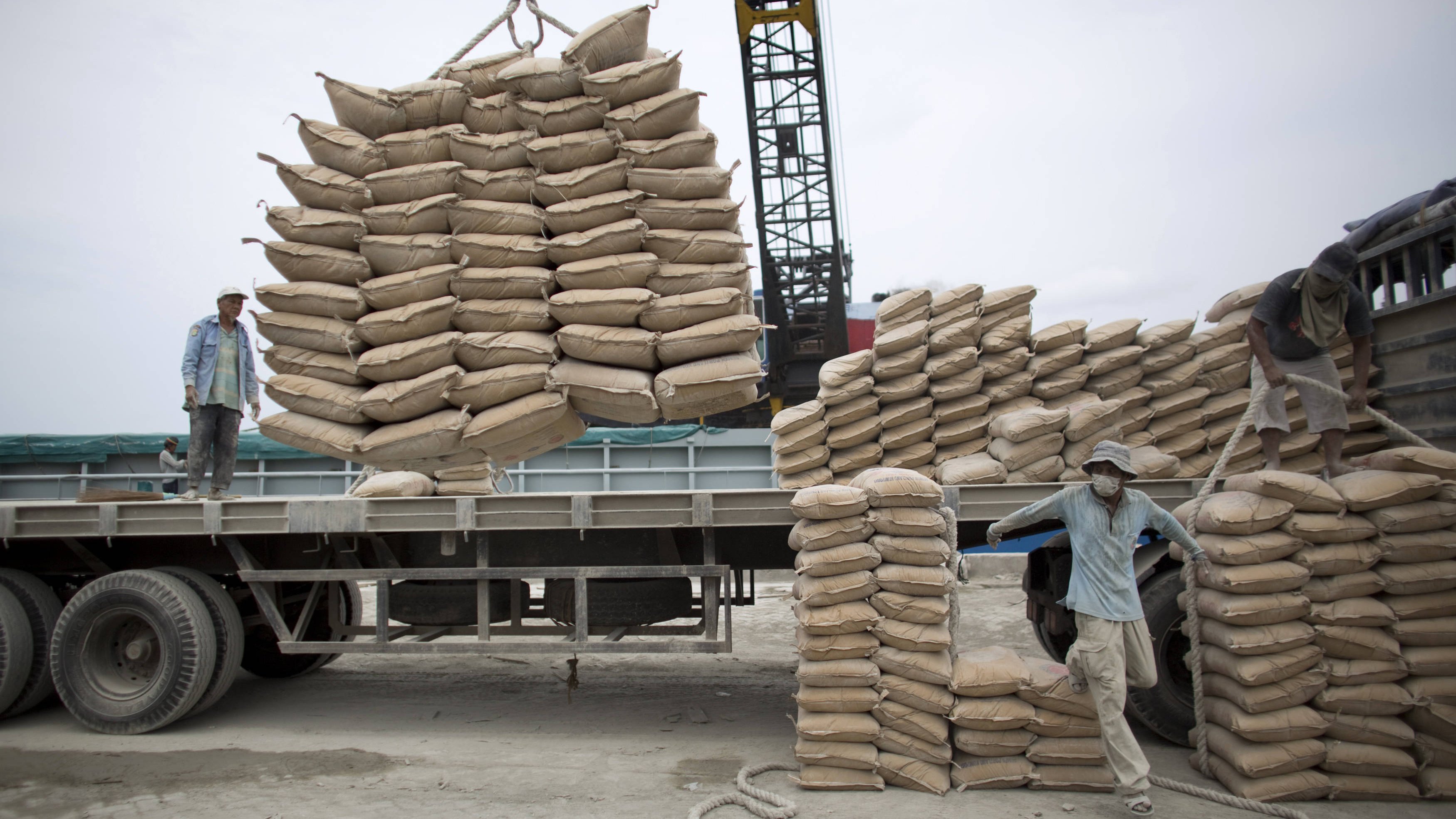 L'image montre une scène de travail au port ou dans un chantier. On y voit des hommes qui manipulent des sacs de sable ou de ciment. Certains sont en train de charger des sacs sur un camion à l'aide d'une grue, tandis que d'autres semblent organiser les sacs déjà empilés. L'environnement est industriel, avec des matériaux de construction et des équipements lourds visibles en arrière-plan. Le ciel est nuageux, ce qui donne une ambiance un peu grise à la scène.