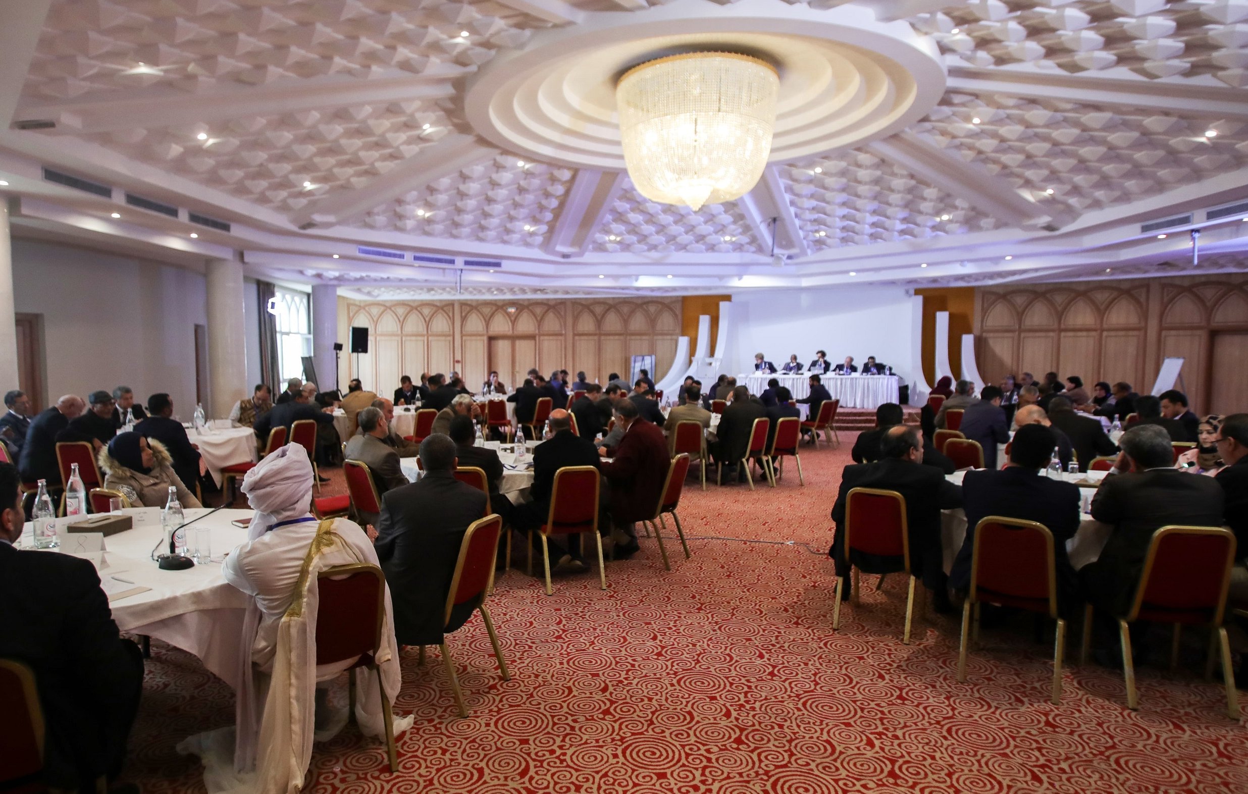 L'image montre une salle de conférence ou de réunion spacieuse, avec de nombreuses personnes assises autour de tables rondes. Les participants semblent engagés dans une discussion ou une présentation, avec un panel sur une estrade à l'avant. La décoration est moderne, avec un plafond orné et un éclairage doux. Les personnes sont habillées de manière formelle, ce qui indique un événement professionnel ou officiel.