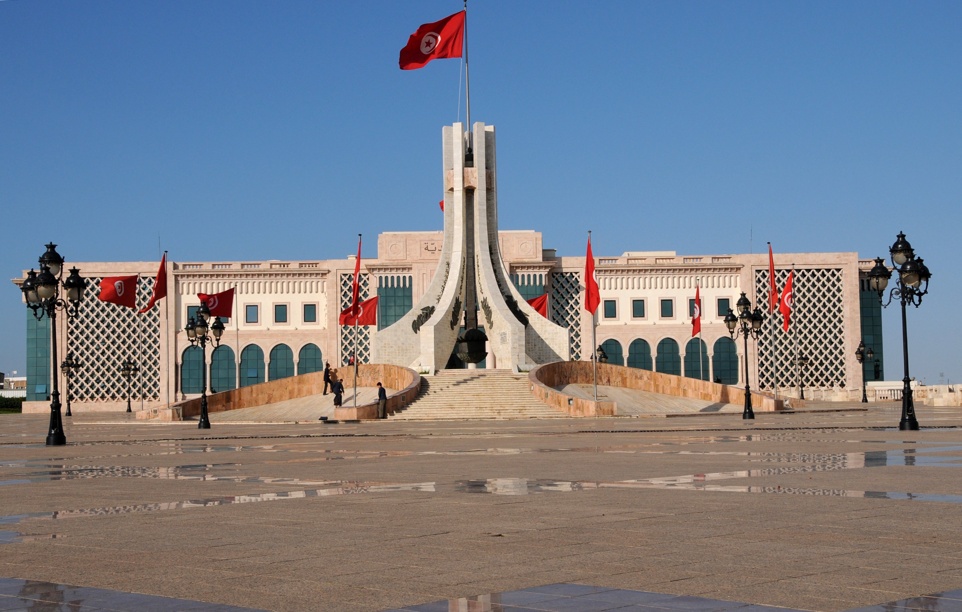 L'image montre un bâtiment imposant avec une architecture distinctive, situé probablement dans un espace public. Au centre, on peut voir un mât portant le drapeau tunisien, avec plusieurs drapeaux rouges autour. L'esplanade devant le bâtiment est pavée, et des lampadaires élégants sont présents sur les côtés. Le ciel est dégagé, ce qui donne une atmosphère lumineuse et claire à la scène. L'architecture semble moderne mais avec des éléments traditionnels.