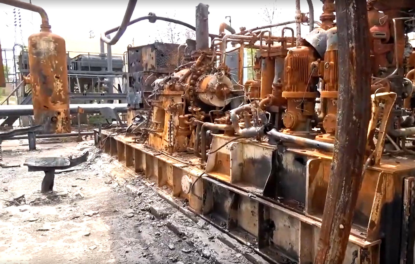 L'image montre une installation industrielle endommagée, probablement suite à un incendie. On peut voir des équipements métalliques, tels que des machines et des tuyaux, recouverts de rouille et de suie. Le sol est noirci par des résidus, indiquant les conséquences d'un sinistre. L'environnement semble très dégradé, avec des éléments de construction en arrière-plan également affectés par des dommages. L'ensemble donne une impression de désolation et de négligence.