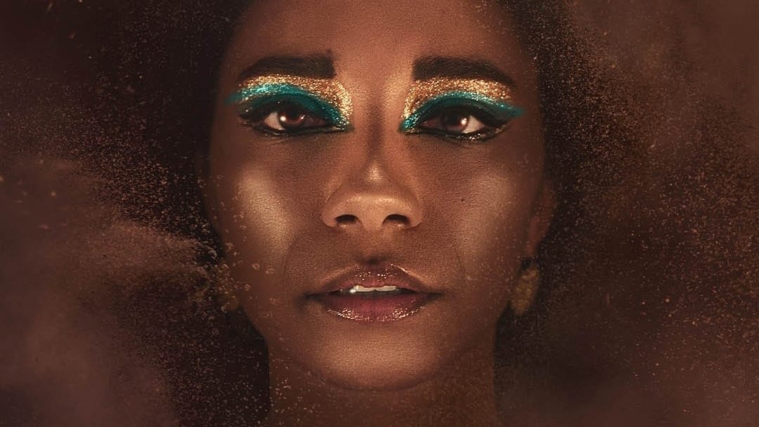 L'image montre une personne au visage ornée, avec un maquillage très artistique mettant en avant des yeux accentués par une ombre à paupières bleu vif. La personne porte une coiffe dorée en forme de serpents, ajoutant à l'aspect majestueux de l'ensemble. Le fond est flou et semble composé de particules dorées, créant une atmosphère mystique et intrigante. L'expression du visage est à la fois sereine et mystérieuse.