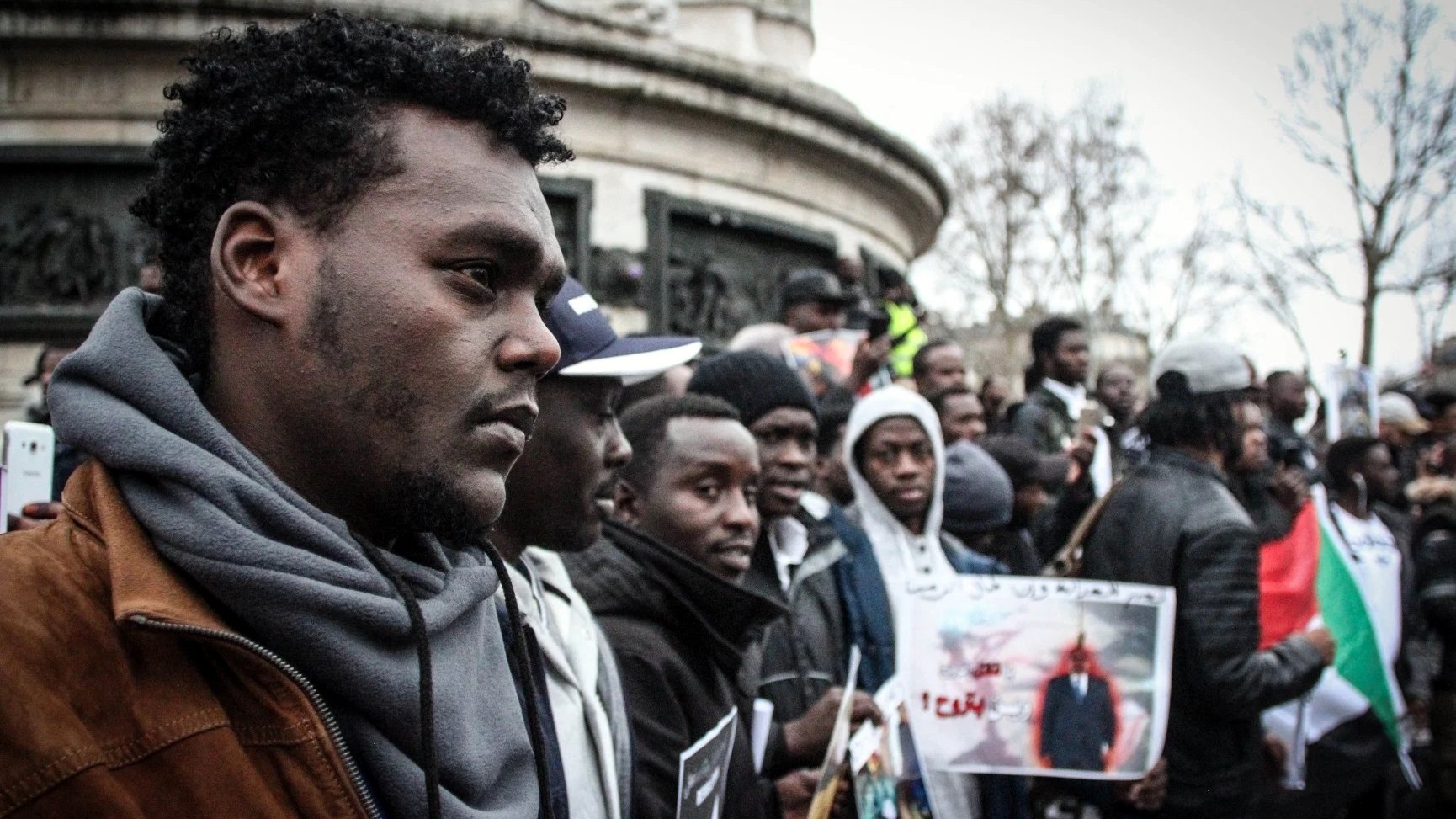 L'image montre un groupe de personnes rassemblées, probablement lors d'une manifestation ou d'un événement public. Au premier plan, un homme au port de tête pensif se distingue, entouré d'autres participants qui tiennent des pancartes et des photos. L'atmosphère semble engagée et solidaire, avec des affiches qui pourraient exprimer des revendications ou des messages de protestation. L'environnement urbain est visible en arrière-plan, suggérant que cet événement se déroule dans un espace public.