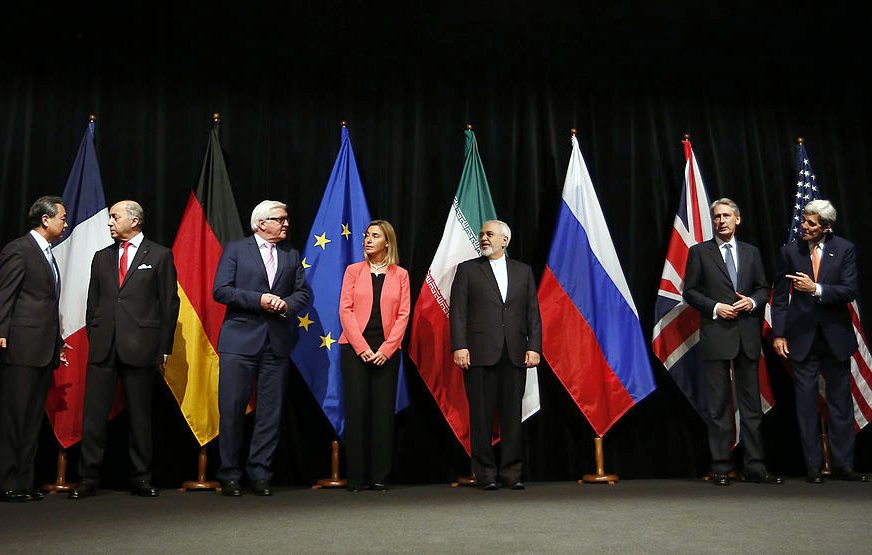 L'image montre une scène officielle avec plusieurs personnes debout en rang, chacune représentant un pays ou une organisation distincte. Derrière eux, il y a des drapeaux de différents pays, notamment la Chine, l'Allemagne, les États-Unis, la Russie et l'Union européenne, entre autres. Les participants semblent engagés dans une discussion ou une négociation, chacun portant un costume formel. L'atmosphère est sérieuse, probablement liée à des pourparlers diplomatiques ou à un sommet international.