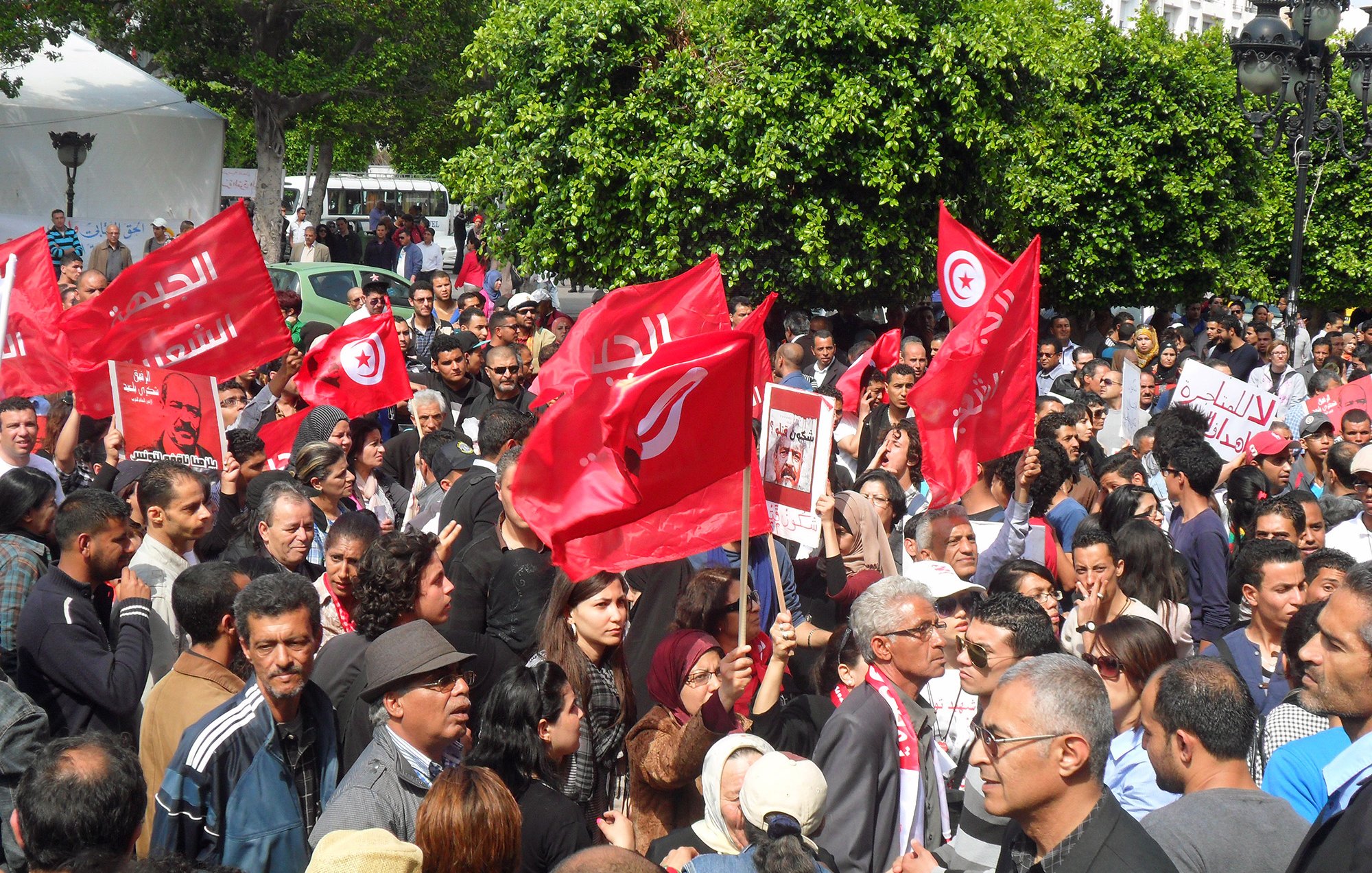 L'image montre une foule rassemblée dans une rue, lors d'une manifestation. Les participants tiennent des drapeaux rouges, probablement portant des symboles ou des slogans liés à la Tunisie. Il y a une grande variété de personnes dans la foule, de différents âges et expressions. L'ambiance semble énergique, avec des signes d'engagement et de participation active. En arrière-plan, on peut voir des bâtiments et des arbres, ce qui indique que l'événement se déroule dans un endroit urbain.