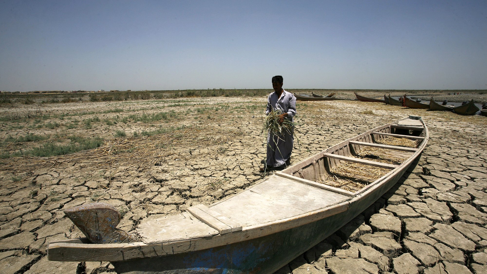 L'image montre un homme debout sur un sol craquelé, typique d'une région aride. Il semble tenir une poignée de végétation, peut-être de l'herbe ou des tiges. À ses pieds se trouve une barque vide, qui est en partie enfouie dans le sol sec. En arrière-plan, on peut apercevoir d'autres barques, mais le paysage aride, sans présence significative d'eau, souligne un environnement désolé et dégradé. Le ciel est dégagé et lumineux, renforçant l'atmosphère de chaleur et de sécheresse.