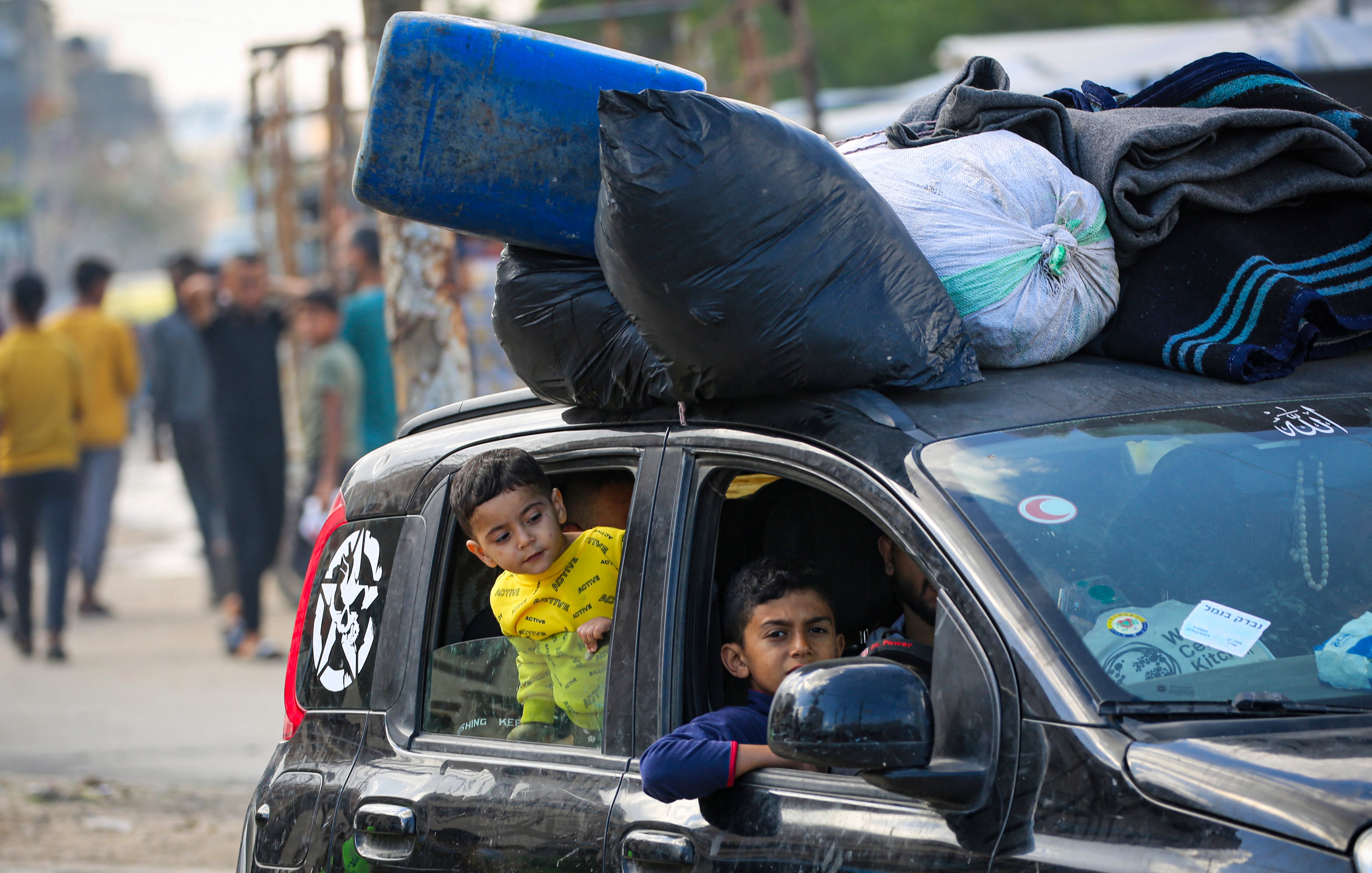Sur cette image, on voit une voiture noire avec plusieurs objets attachés sur le toit, notamment des sacs et un bidon bleu. À l'intérieur du véhicule, il y a deux enfants. L'un des enfants, portant un vêtement jaune avec un imprimé, regarde par la fenêtre de la voiture. L'autre enfant, assis à l'avant, regarde également dehors. En arrière-plan, il y a plusieurs personnes qui semblent se déplacer, ainsi qu'un environnement urbain sans détails précis.