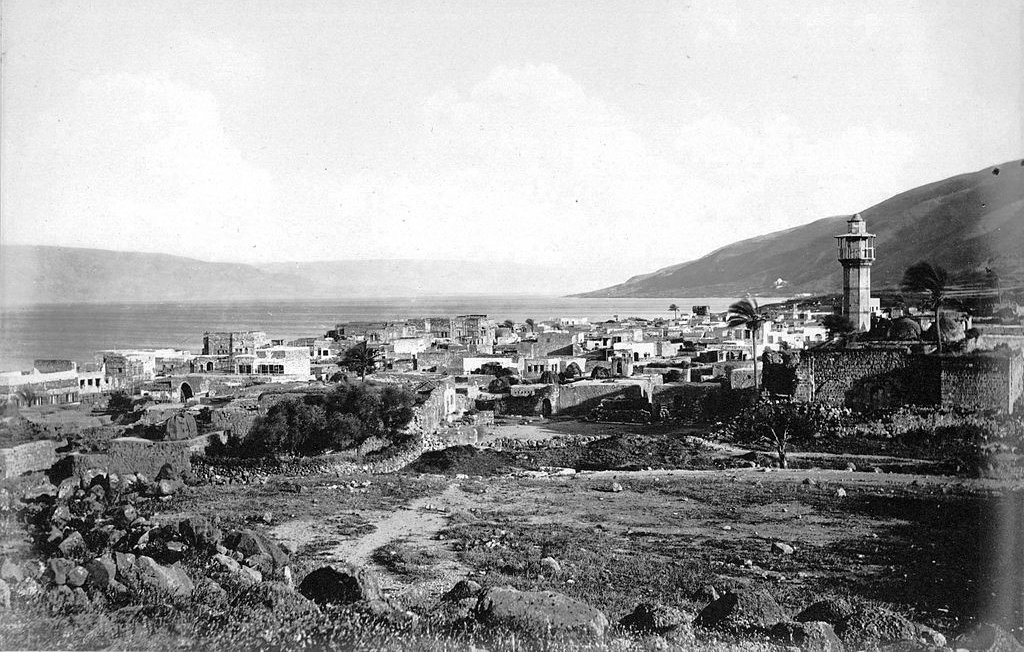 L'image montre un paysage historique d'un village côtier, probablement situé près d'un lac ou d'une mer, avec des bâtiments disposés le long de la promenade. Il y a un minaret ou une tour surmontée d'un dôme, indiquant la présence d'une culture musulmane. En arrière-plan, on peut voir des collines qui encadrent le paysage. Le ciel est légèrement nuageux, créant une ambiance tranquille. L'ensemble de l'image a un aspect noir et blanc, ce qui lui donne un caractère ancien.