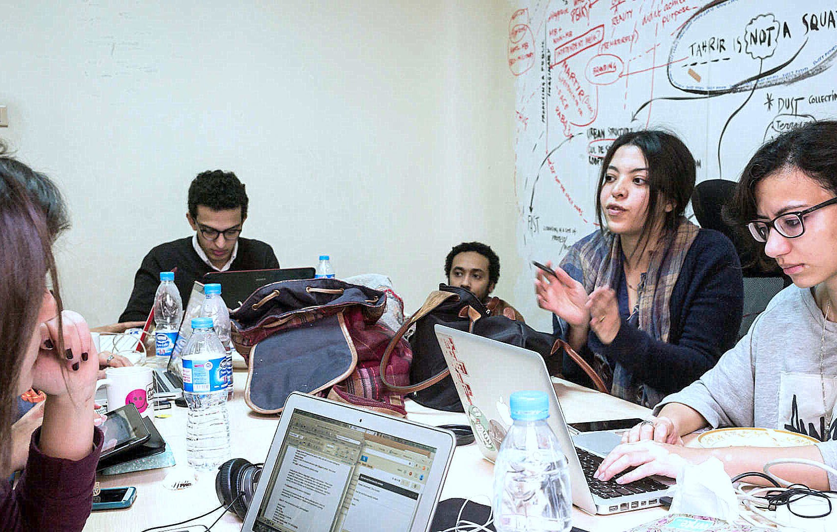 L'image montre un groupe de personnes assises autour d'une table, travaillant sur des ordinateurs portables. Il y a des bouteilles d'eau et des sacs dispersés sur la table. Une femme semble expliquer quelque chose avec enthousiasme, tandis que les autres écoutent attentivement. Le mur derrière eux est couvert de notes et de schémas, suggérant une ambiance créative et collaborative.