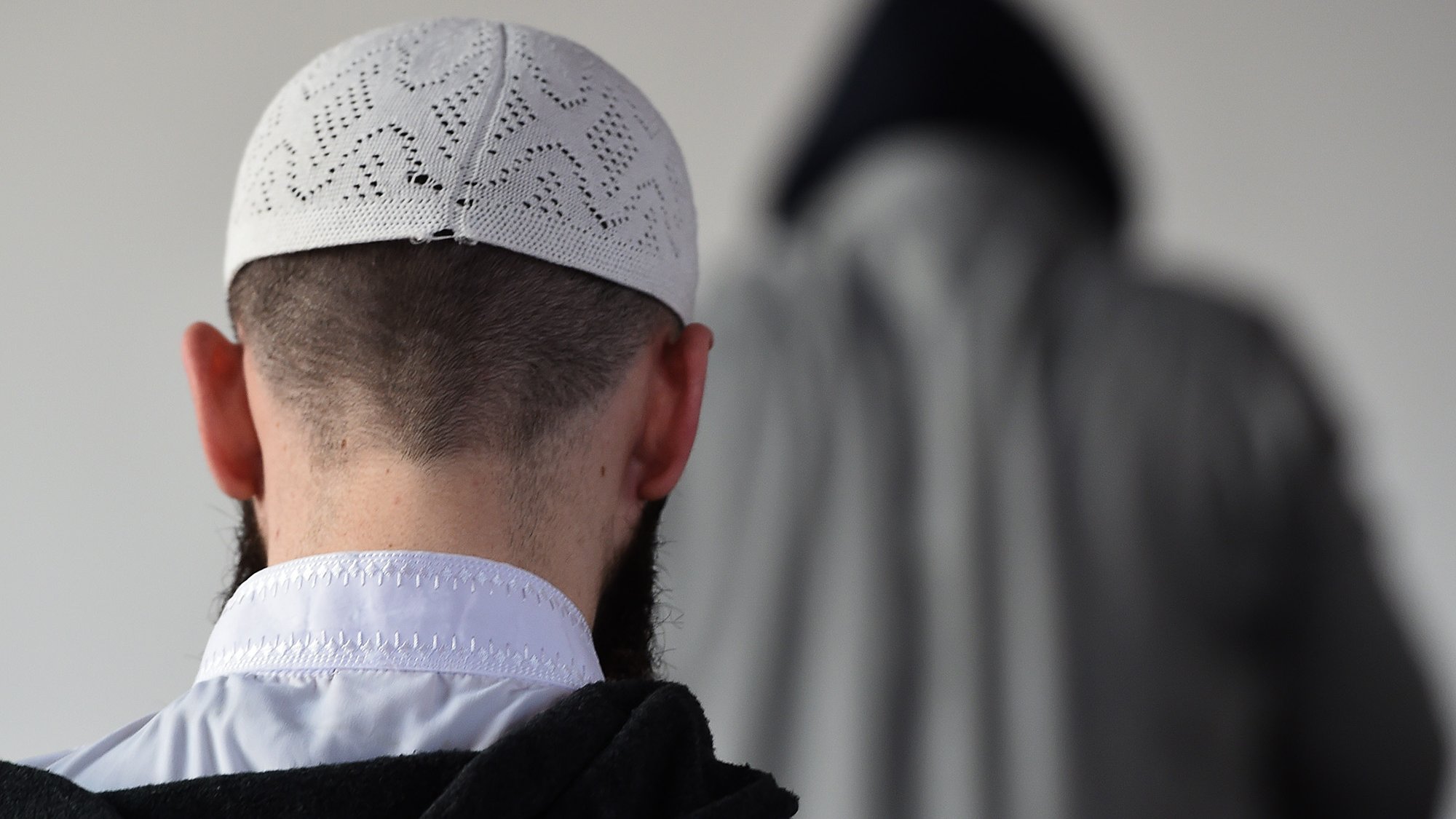 L'image montre un homme de dos, portant un taqiyah (un couvre-chef traditionnel), avec une barbe visible. En arrière-plan, on peut apercevoir une silhouette floue d'une autre personne, vêtue d'une cape ou d'un vêtement similaire. L'environnement semble simple et neutre, avec peu d'éléments distrayants. La scène évoque une ambiance calme et contemplative.
