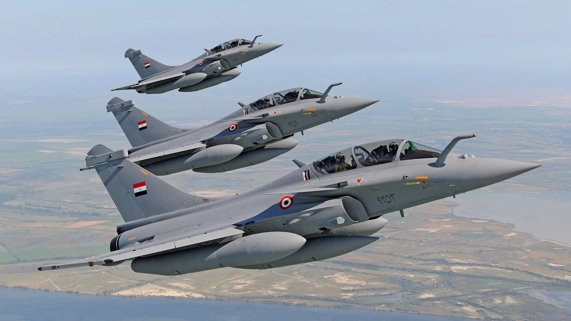 L'image montre un groupe de jets de chasse en plein vol. Trois avions de chasse, probablement des Rafales, sont alignés en formation, avec des couleurs principalement grises. Chaque avion porte des marqueurs et des emblèmes, semblant appartenir à une force aérienne. Le décor en arrière-plan indique un paysage vaste, avec des zones de terre et de l'eau visible, suggérant un vol à basse altitude. L'ensemble dégage une impression de puissance et de précision militaire.