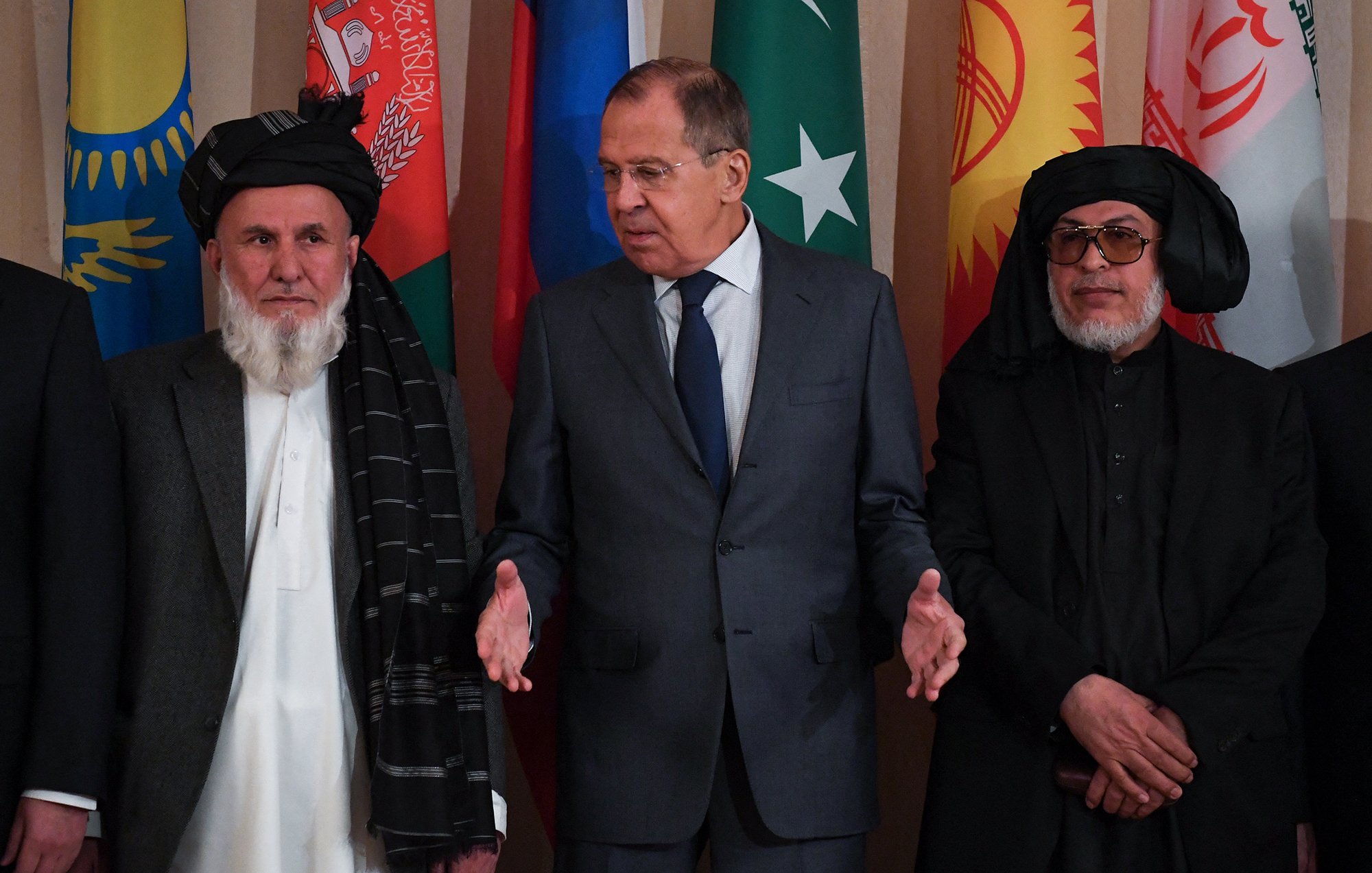 L'image montre un groupe d'hommes lors d'un événement officiel. Au centre, un homme en costume cravate semble s'adresser aux autres, tandis que ceux à gauche et à droite portent des vêtements traditionnels. En arrière-plan, des drapeaux de différents pays sont affichés, ce qui suggère un contexte diplomatique ou de négociation internationale. L'atmosphère paraît formelle et engagée.