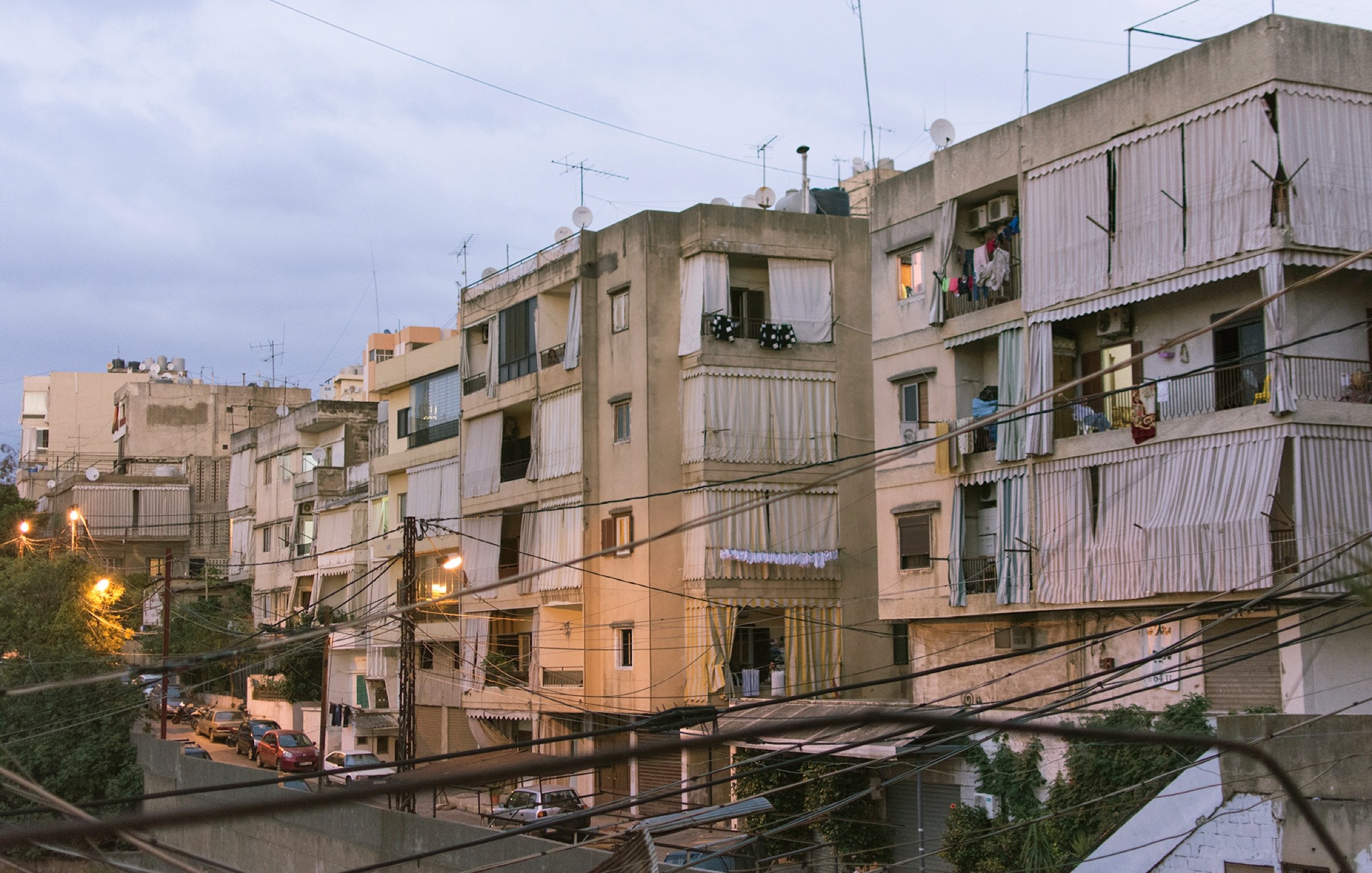 L'image montre un ensemble d'immeubles résidentiels dans une zone urbaine. Les bâtiments semblent anciens et sont de plusieurs étages, avec des balcons où l'on peut voir des vêtements étendus. La lumière du soir crée une atmosphère douce, et des fils électriques traversent l'image. On aperçoit également des voitures garées le long de la rue en contrebas. L'ensemble donne une impression de vie citadine typique, avec un mélange de modernité et de caractère ancien.