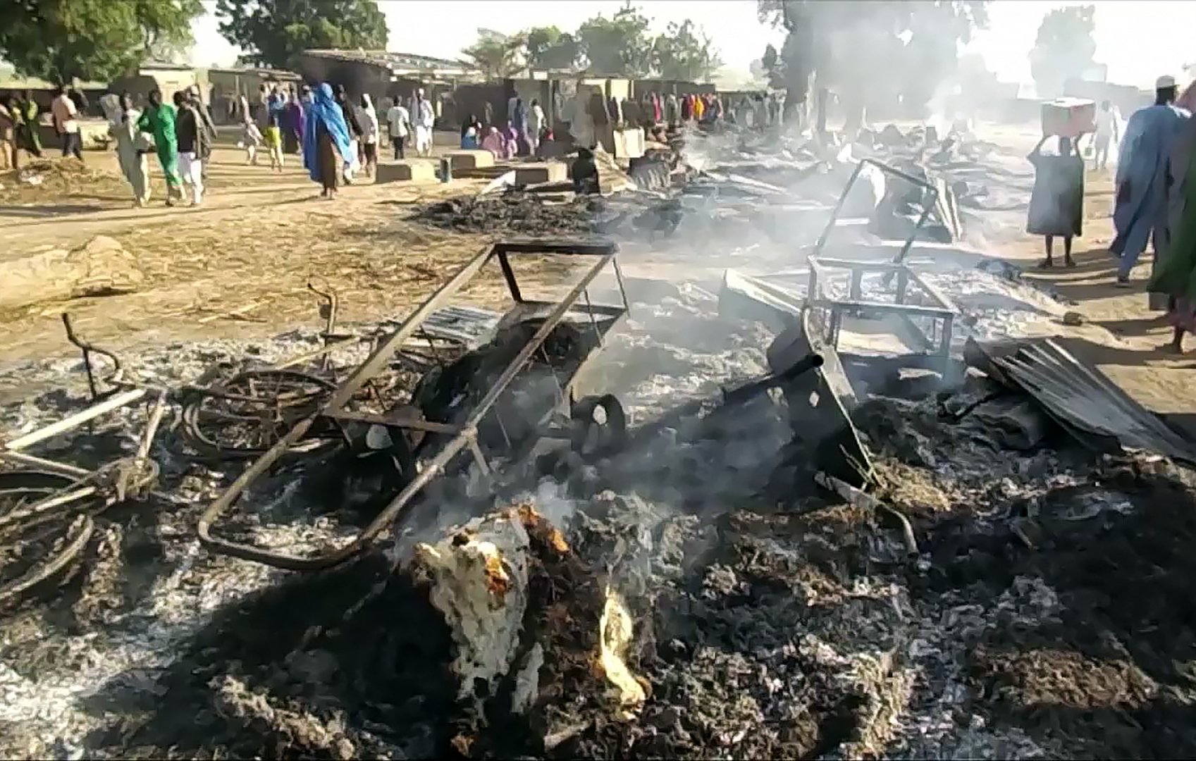 L'image montre une scène de destruction, où des débris brûlés et des objets en métal tordu sont dispersés sur le sol. De la fumée s'élève de restes calcinés, suggérant qu'un incendie a eu lieu récemment. À l'arrière-plan, on aperçoit des personnes en train de se déplacer, sur un terrain qui semble être une communauté ou un marché. L'atmosphère est sombre et le paysage est marqué par les dégâts causés par le feu.