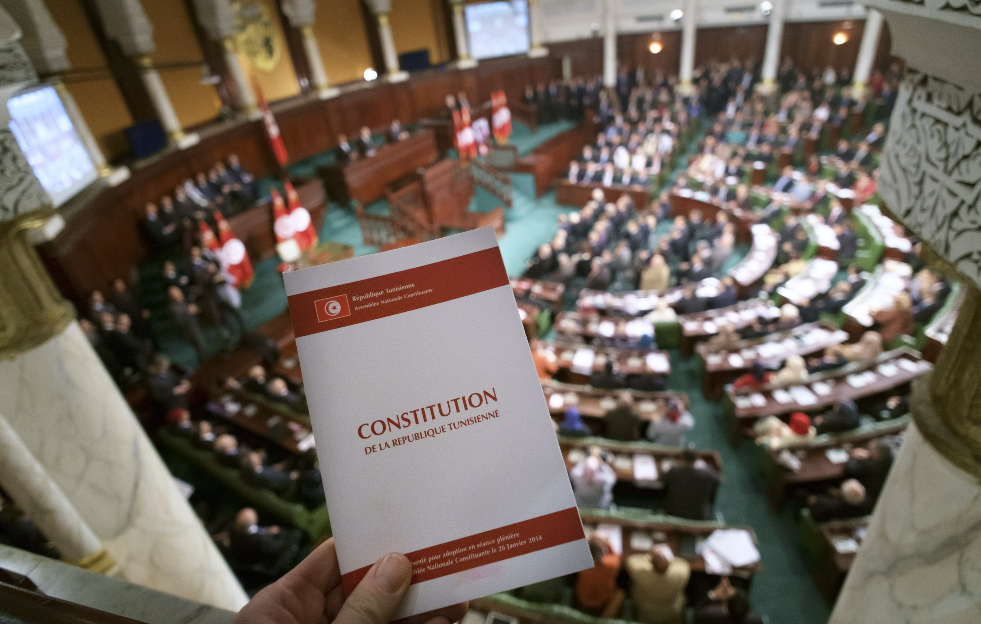 L'image montre une scène à l'intérieur d'une assemblée, probablement un parlement, avec un grand nombre de personnes assises dans une salle. Au premier plan, on voit une personne tenant un document portant le titre "CONSTITUTION DE LA RÉPUBLIQUE TUNISIENNE". L'assemblée est ornée de drapeaux, et l'ambiance semble solennelle, suggérant un moment important lié à la constitution ou à la législation.