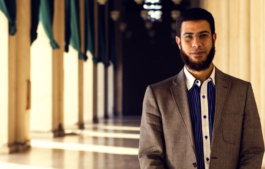 L'image montre un homme portant un costume avec une chemise à rayures. Il a une barbe bien entretenue et porte des lunettes. En arrière-plan, on peut voir un couloir avec des colonnes et des jeux d'ombres créés par la lumière qui entre. L'ambiance semble calme et chaleureuse, avec des tentures vertes accentuant l'architecture.