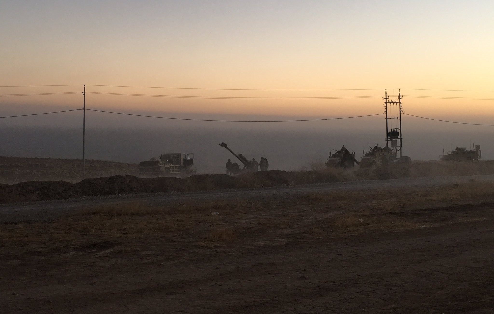 L'image montre un paysage de chantier au crépuscule, avec un ciel teinté de couleurs douces allant du jaune à l'orange. On aperçoit des machines de construction et des travailleurs en silhouettes, entourés de poussière. À l'arrière-plan, on distingue des poteaux électriques. Le sol semble sec et rocailleux, suggérant une zone de travaux en cours. L'atmosphère est calme, mais il y a une impression d'activité et d'effort en cours.