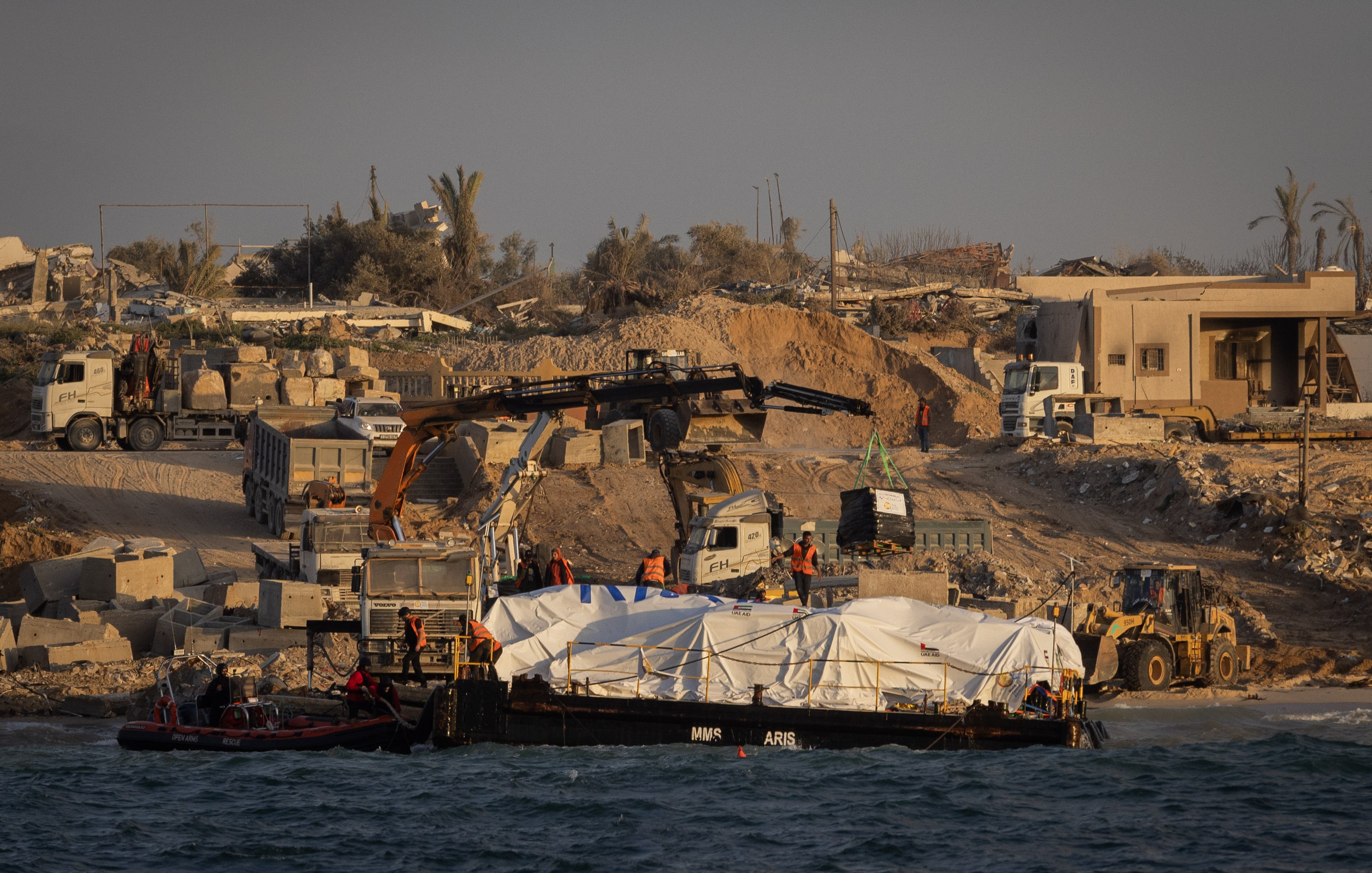 L'image montre une scène de construction ou de travaux sur une côte. On peut voir un bateau sur l'eau avec une grande bâche blanche, probablement transportant des matériaux ou des fournitures. Sur la rive, il y a des machines de construction, comme des pelleteuses et des camions, ainsi que des travailleurs en action. En arrière-plan, des bâtiments ruinés et des palmiers suggèrent un environnement en mutation, peut-être en raison de travaux de développement ou de reconstruction. L'atmosphère générale semble celle d'un chantier en cours.
