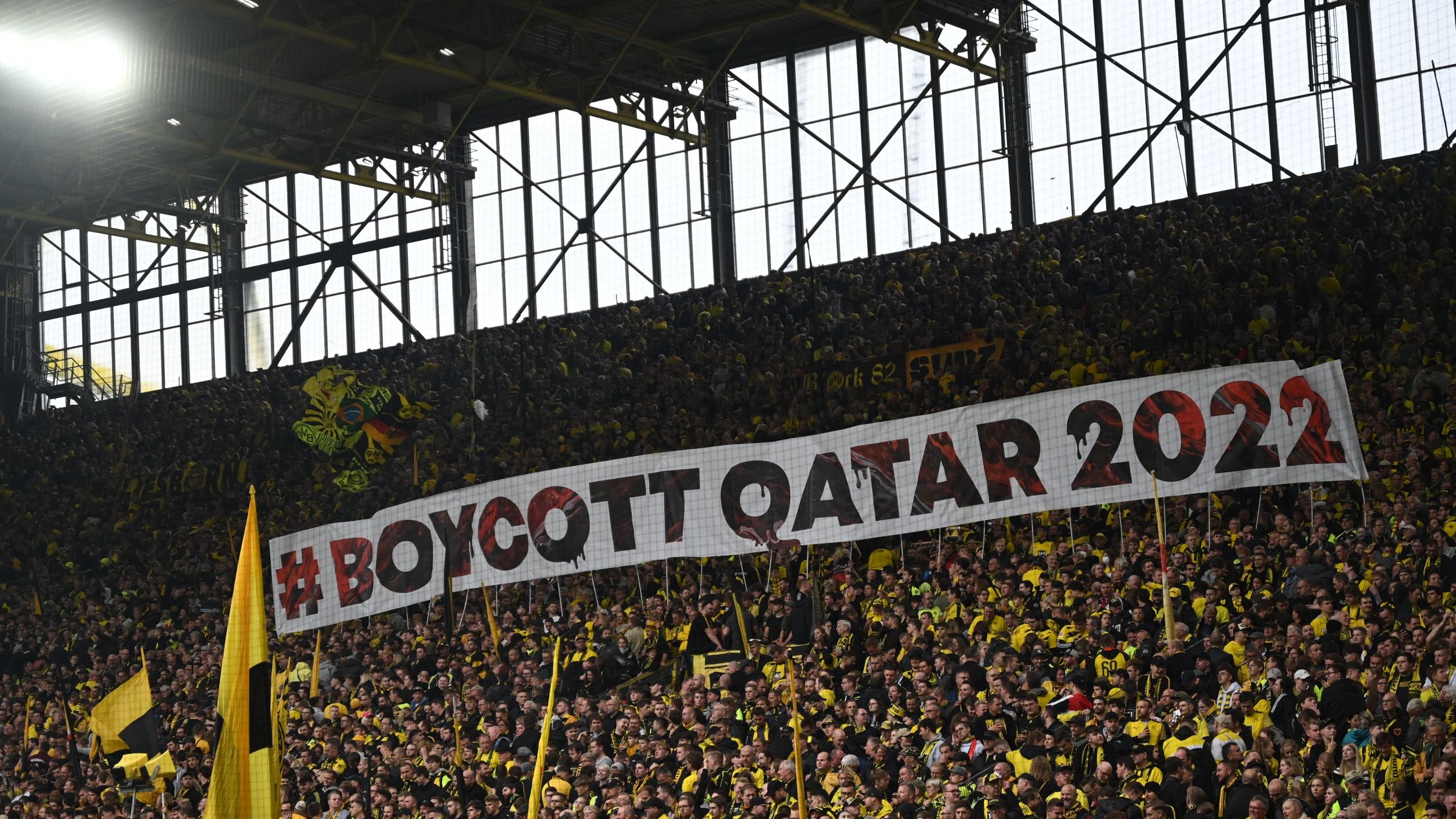 L'image montre un grand groupe de supporters dans un stade, portant des vêtements jaunes et noirs. Ils tiennent une immense banderole sur laquelle est inscrit "#BOYCOTT QATAR 2022". L'atmosphère semble dynamique et engagée, avec un fort sentiment de solidarité parmi les fans. Les tribunes sont pleines, créant une ambiance électrisante.