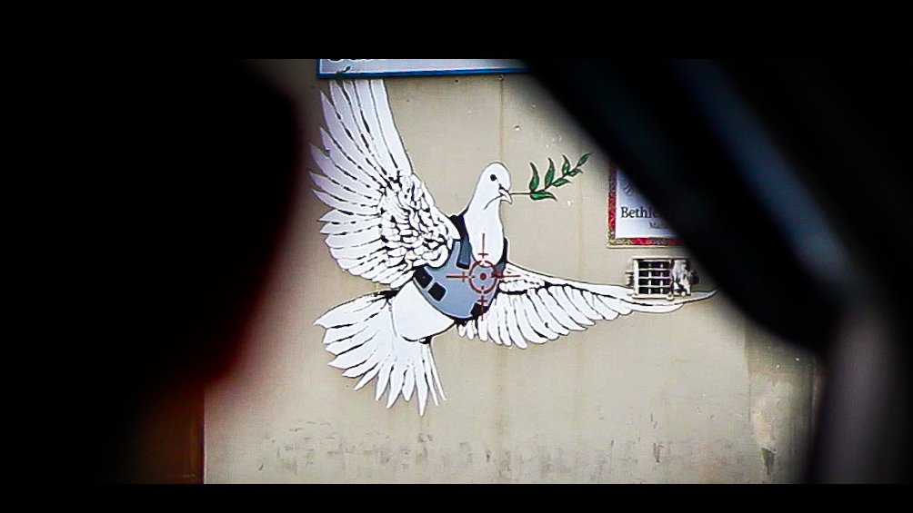 L'image montre une fresque murale représentant une colombe aux grandes ailes déployées. La colombe tient une branche d'olivier dans son bec, symbole de paix. Elle semble être stylisée avec des éléments modernes, comme un gilet pare-balles, ce qui pourrait faire allusion à des thèmes de conflits ou de sécurité. Le fond est neutre et sans éléments distracteurs, mettant en valeur la colombe et son message symbolique.