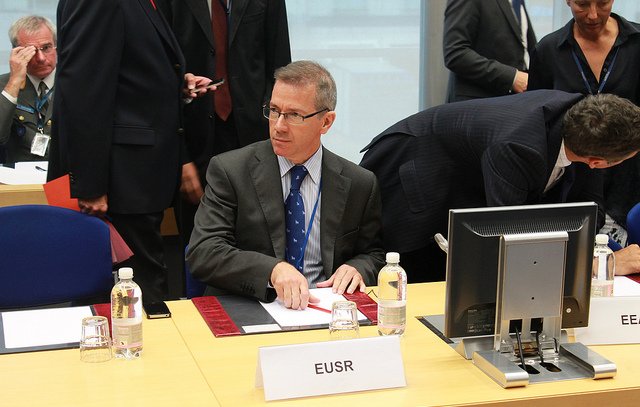 L'image montre une scène de réunion où plusieurs personnes sont présentes autour d'une table. Un homme, assis au premier plan, porte un costume gris et une cravate bleue à pois. Il semble concentré, en train d'organiser des documents devant lui. Sur la table, il y a des bouteilles d'eau et des documents disposés. À l'arrière-plan, d'autres participants sont visibles, certains discutant ou se déplaçant. L'environnement semble formel, typique d'une conférence ou d'une réunion professionnelle.