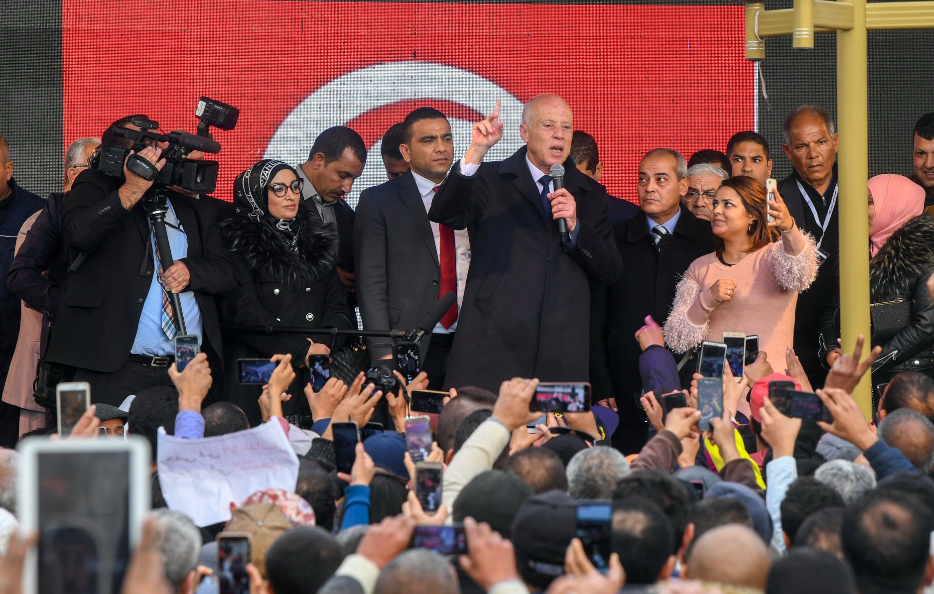 L'image montre un groupe de personnes rassemblées devant une scène où un homme, probablement un leader politique, prend la parole. Il est entouré de plusieurs personnes, dont des membres de la sécurité et des journalistes avec des caméras. La foule est très engagée, beaucoup de gens tiennent des téléphones pour filmer ou prendre des photos. L'arrière-plan présente des affiches ou des bannières, probablement en rapport avec un événement important. L'ambiance semble dynamique et pleine d'énergie.