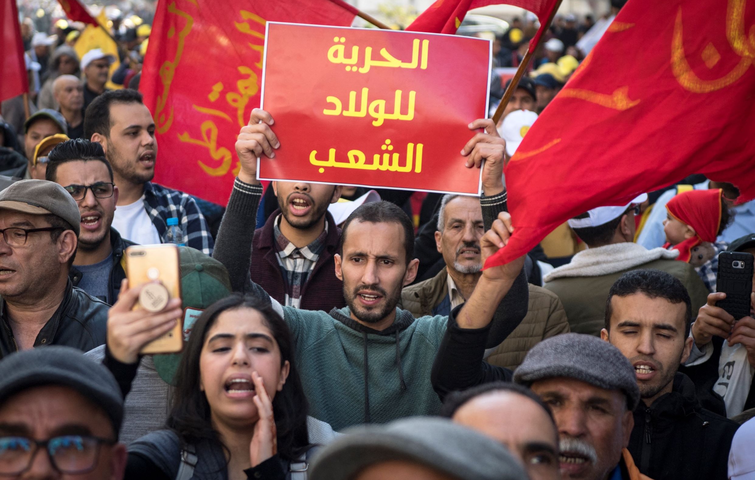 L'image montre une foule de manifestants, brandissant des drapeaux rouges. Au centre, un homme tient une pancarte avec le texte en arabe, tandis que d'autres personnes autour de lui semblent engagées dans la manifestation. On peut ressentir une atmosphère de protestation avec des expressions de détermination sur les visages des participants.