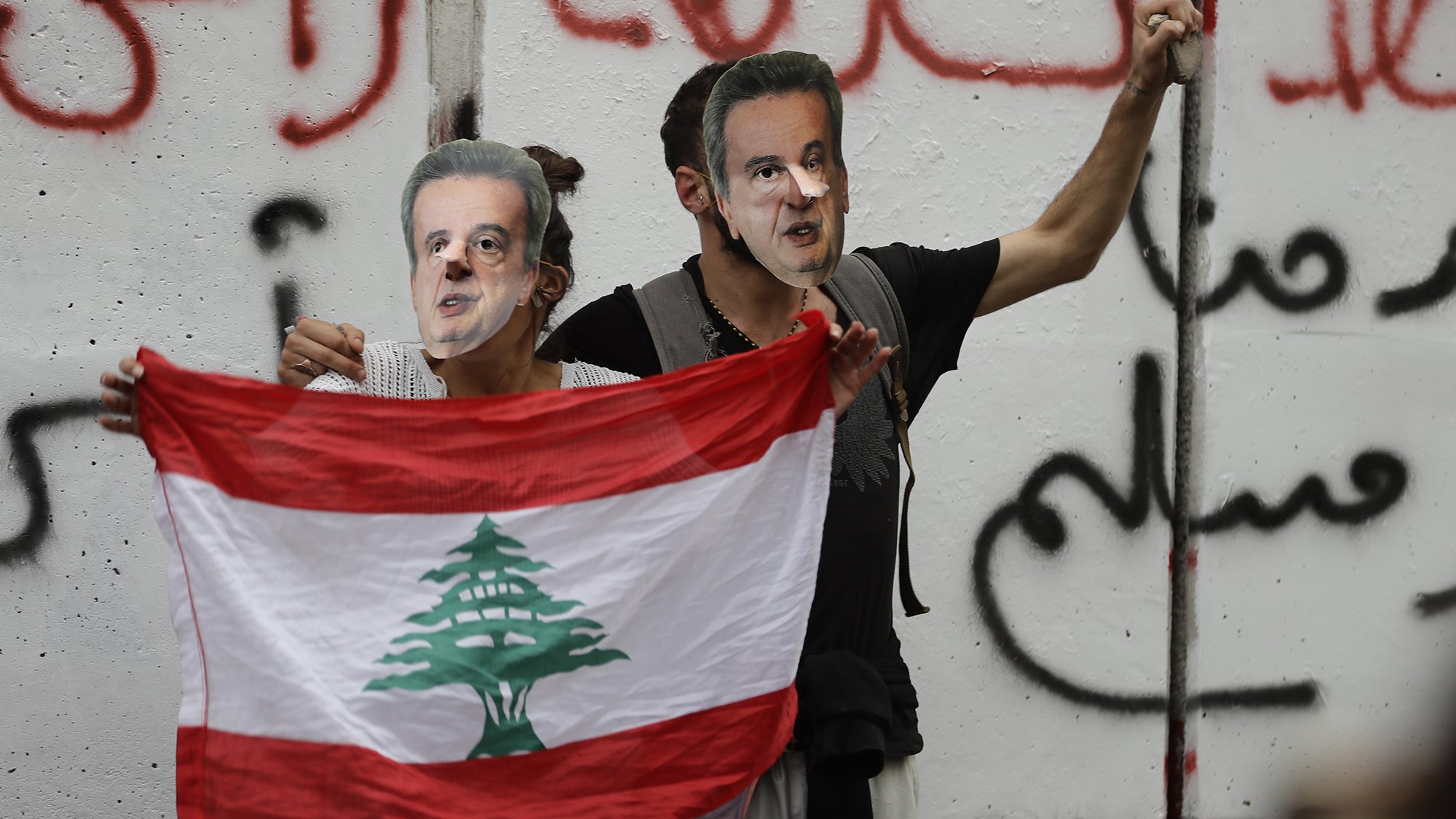 L'image montre deux personnes tenant un drapeau libanais tout en portant des masques représentant un homme. Ils se tiennent devant un mur avec des inscriptions en arabe, probablement dans un contexte de manifestation ou de protestation. L'ambiance semble chargée d'émotion et de revendications politiques.