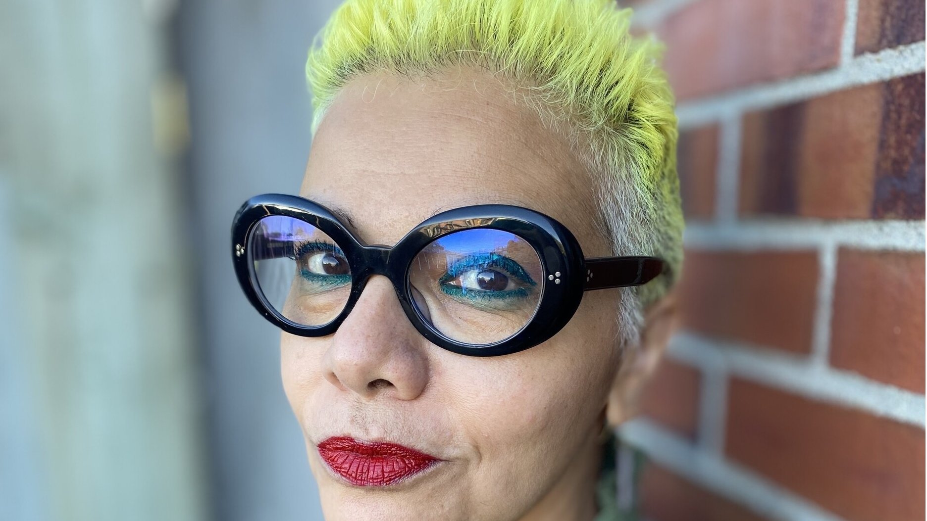 L'image montre une personne avec des cheveux très courts teints en jaune vif. Elle porte de grandes lunettes noires au style distinctif. Son maquillage comprend des lèvres rouge vif. Le fond présente un mur en briques, ce qui ajoute un contraste avec son look. Elle a une expression confiante et souriante.