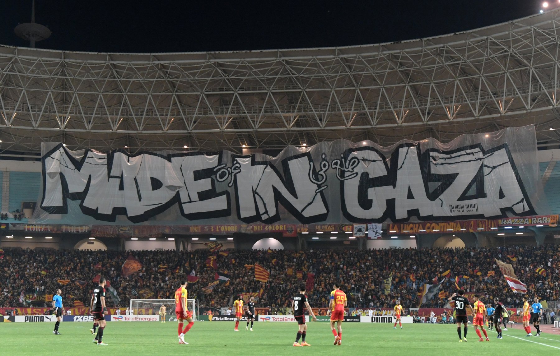 L'image montre un stade de football avec une grande banderole affichée. La banderole est ornée du texte "MADE IN GAZA" écrit en lettres stylisées et imposantes. Le stade est rempli de supporters, et l'atmosphère semble animée. On peut voir des joueurs sur le terrain, ce qui indique qu'un match est en cours. L'ambiance est festive et engagée.