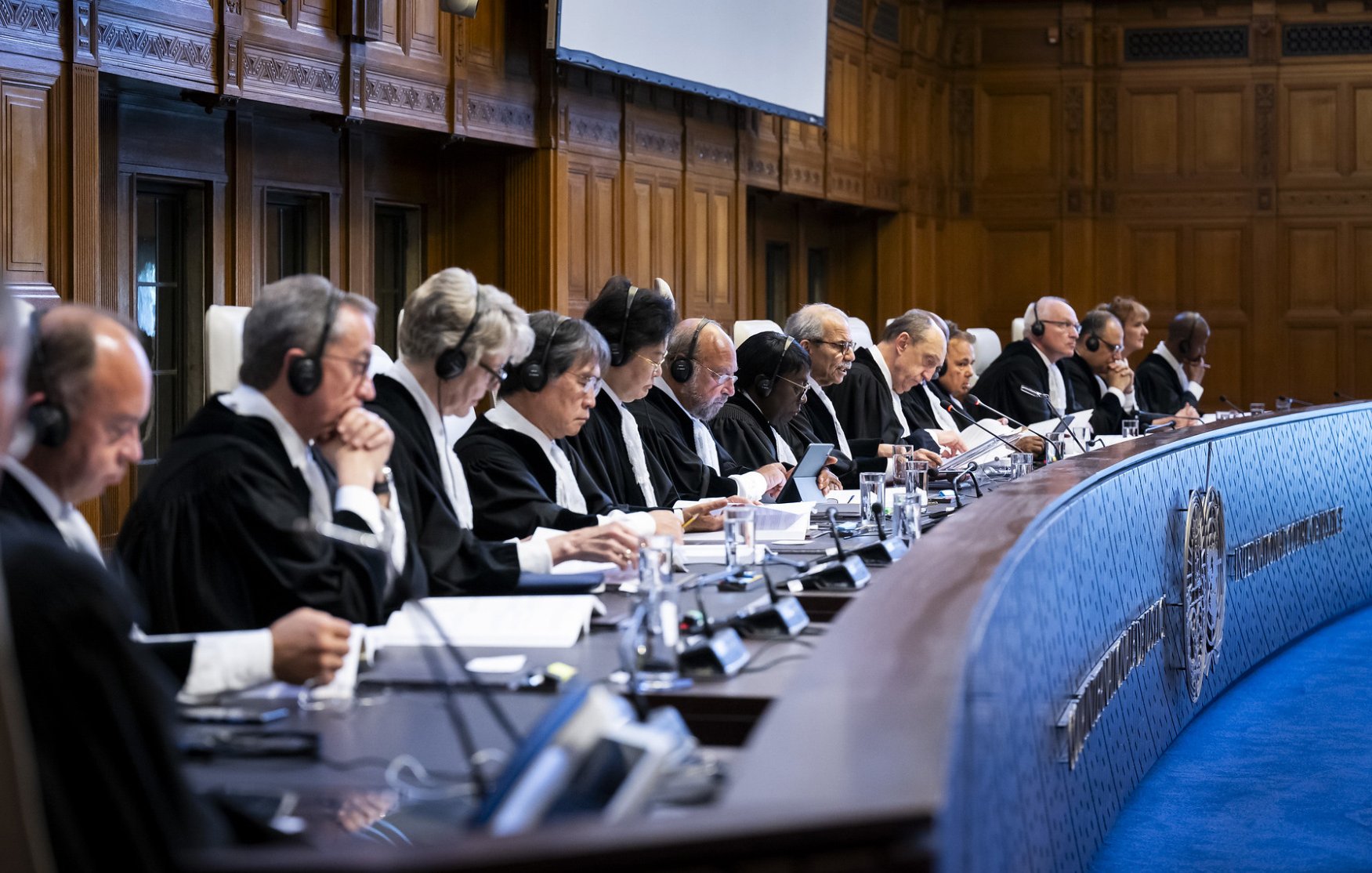 L'image montre une salle de tribunal où plusieurs juges sont assis en ligne dans un espace formel et bien éclairé. Ils portent des robes noires et des écouteurs, symbolisant une séance judiciaire importante. Le décor est élégant, avec des murs en bois et un grand écran au fond. Les juges semblent concentrés sur les documents devant eux, participant à une discussion ou à une audience. L'atmosphère est sérieuse, en accord avec la nature de leurs fonctions.